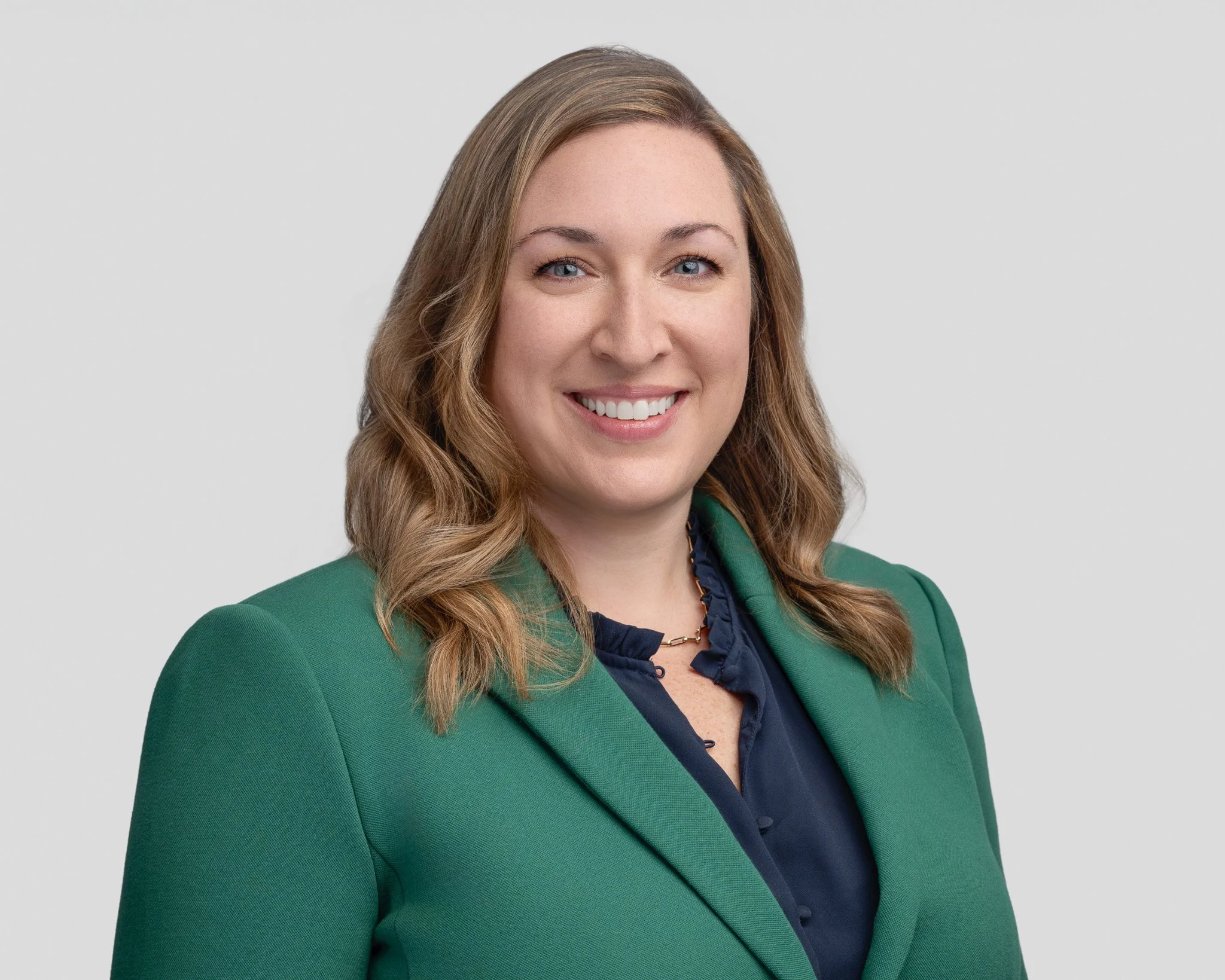 A professional woman with shoulder-length wavy blonde hair, wearing a green blazer over a dark blue blouse, smiling against a light gray background.