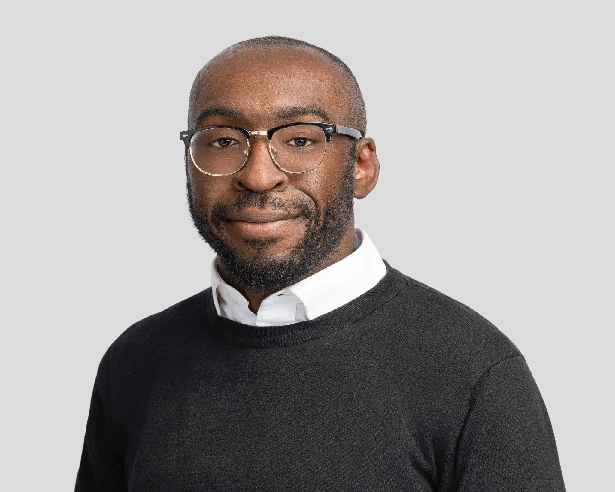 A man with dark skin, glasses, a beard, and short hair wearing a white collared shirt under a black sweater posing against a plain light gray background.
