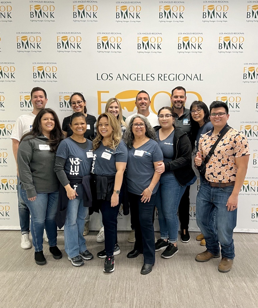 A group of twelve diverse people standing together in front of a backdrop that reads 'Los Angeles Regional Food Bank' and shows its logo. They are smiling and appear to be at an event or gathering, with some wearing matching shirts.