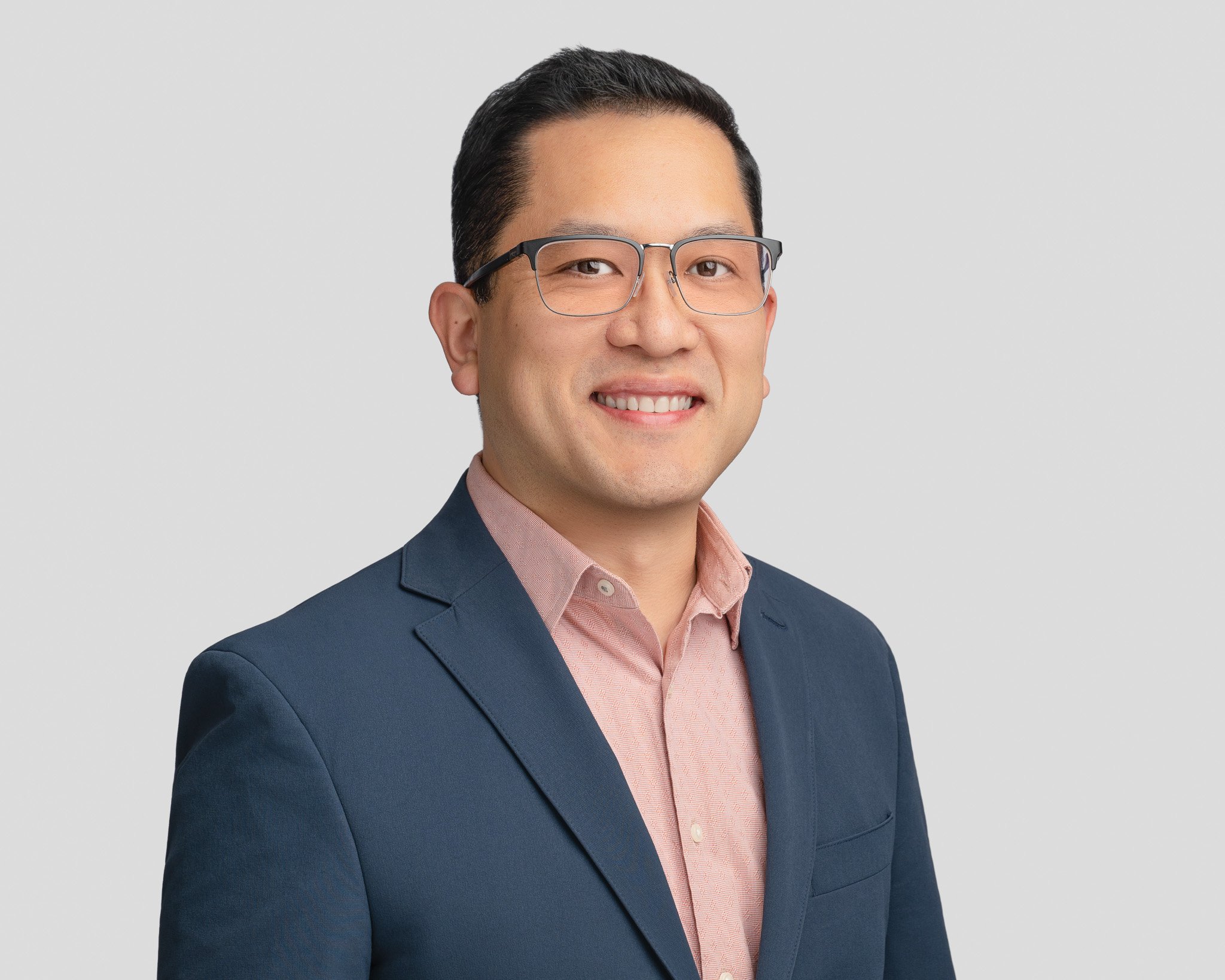 Professional headshot of a man wearing glasses, a pink dress shirt, and a navy blazer, smiling against a plain light gray background.