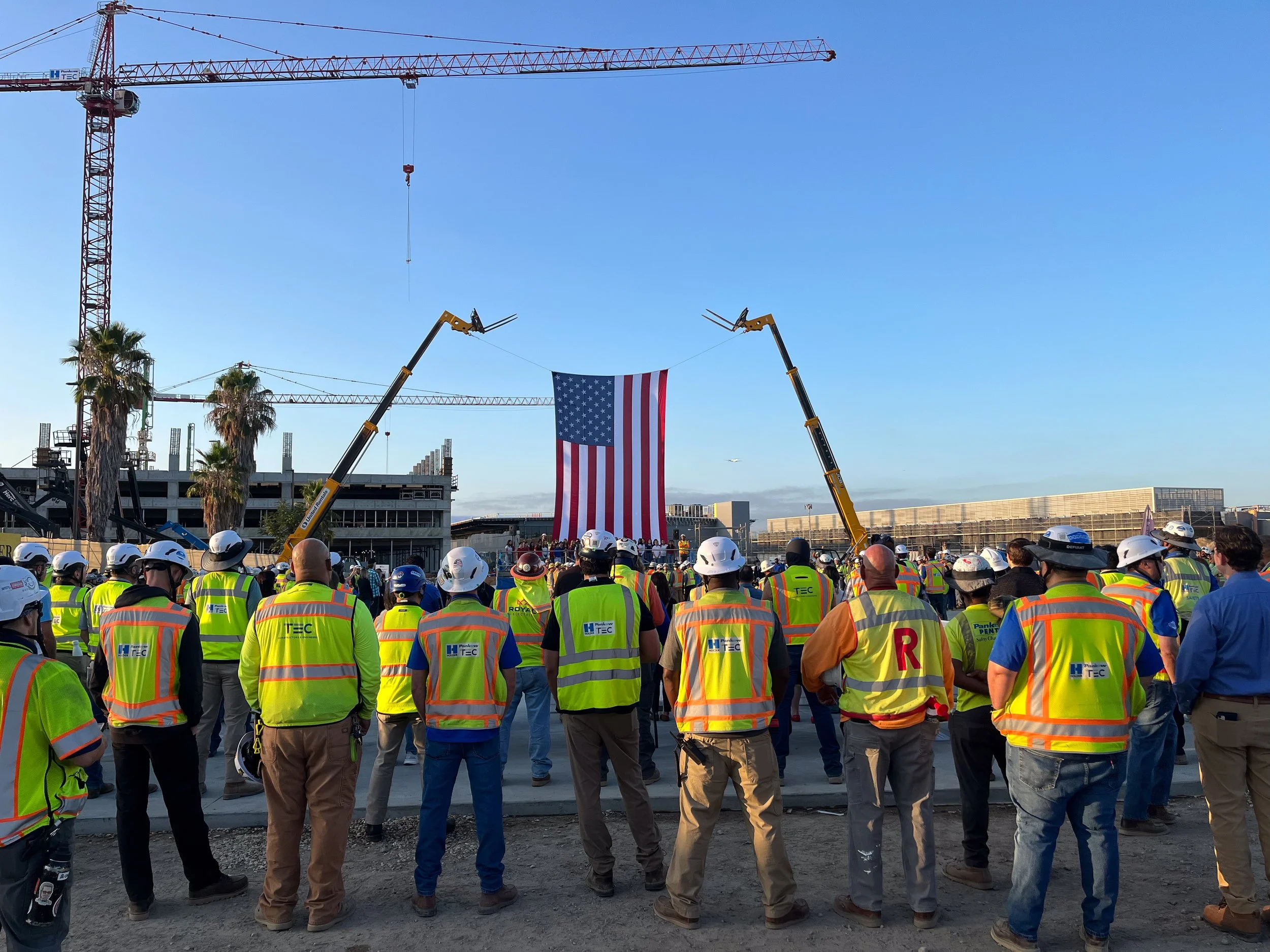 Construction workers and officials gathered outdoors to unveil an American flag. Two cranes lift the flag as people watch, with a clear blue sky above.