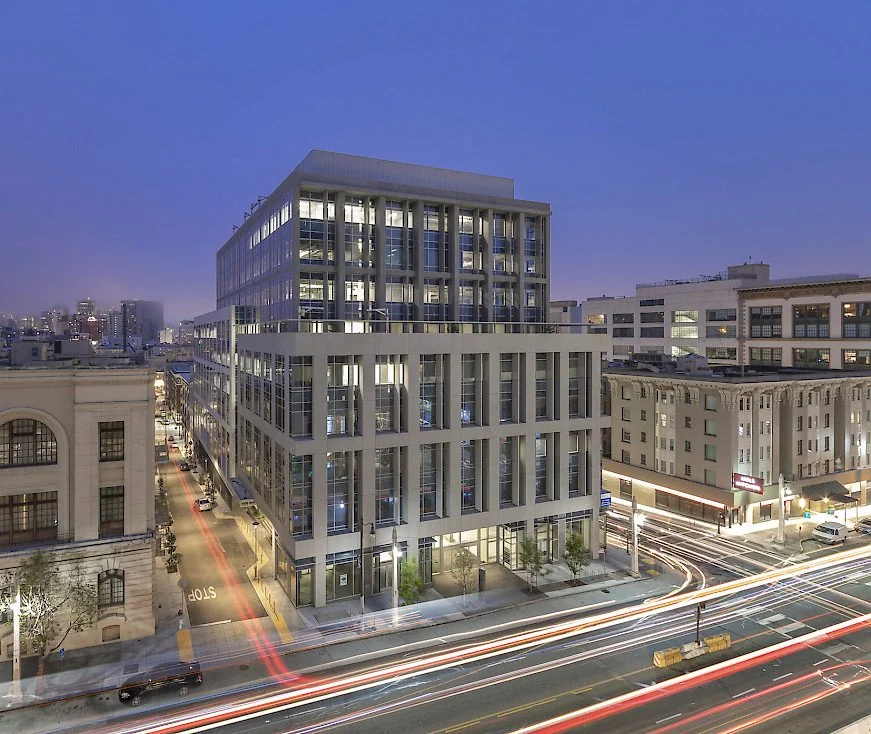 Nighttime cityscape featuring a modern multi-story building with glass windows and vertical concrete beams, surrounded by older buildings, with light trails from moving vehicles on the street.