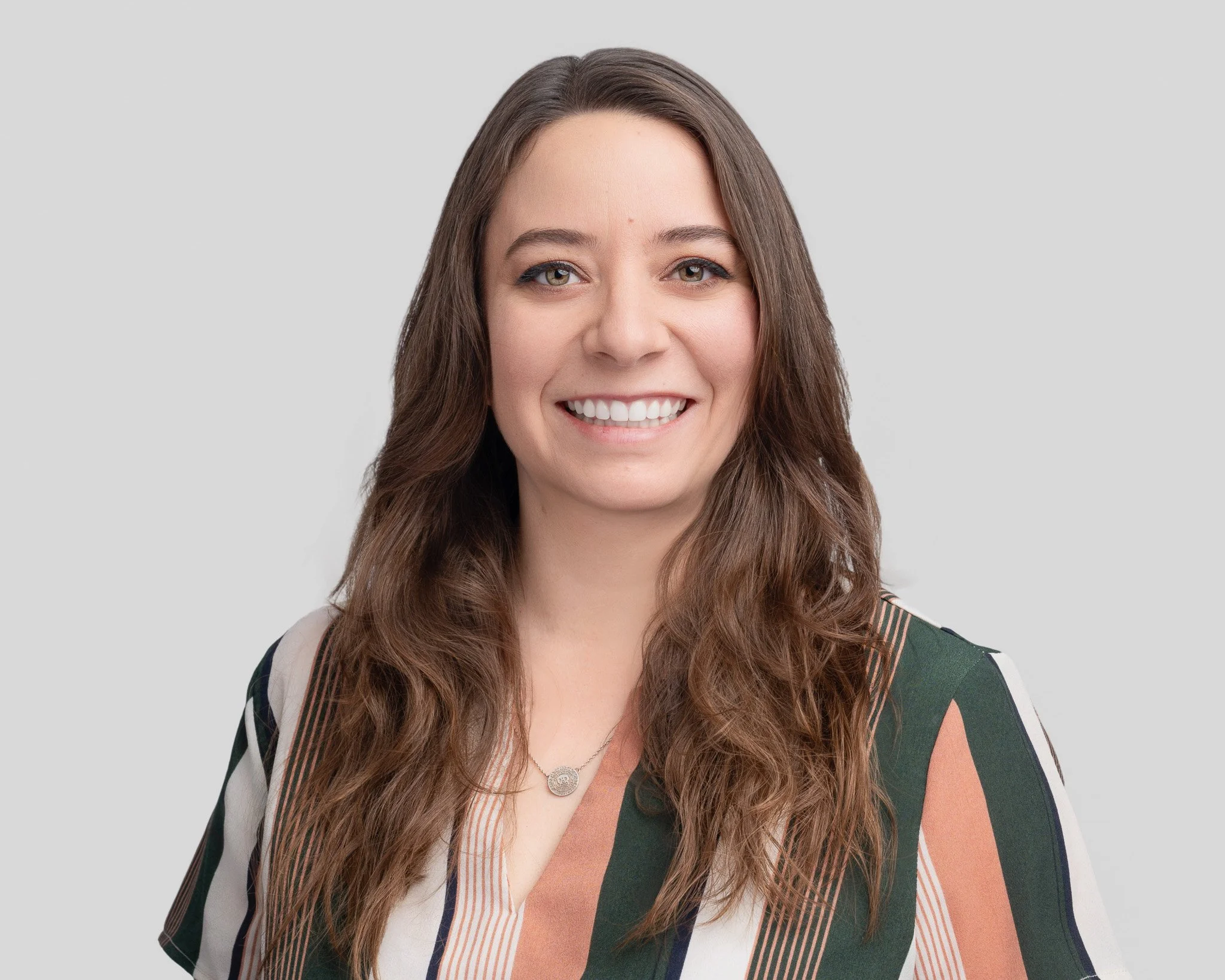 A woman with long, wavy brown hair, smiling, wearing a multicolored striped blouse and a necklace, against a light gray background.