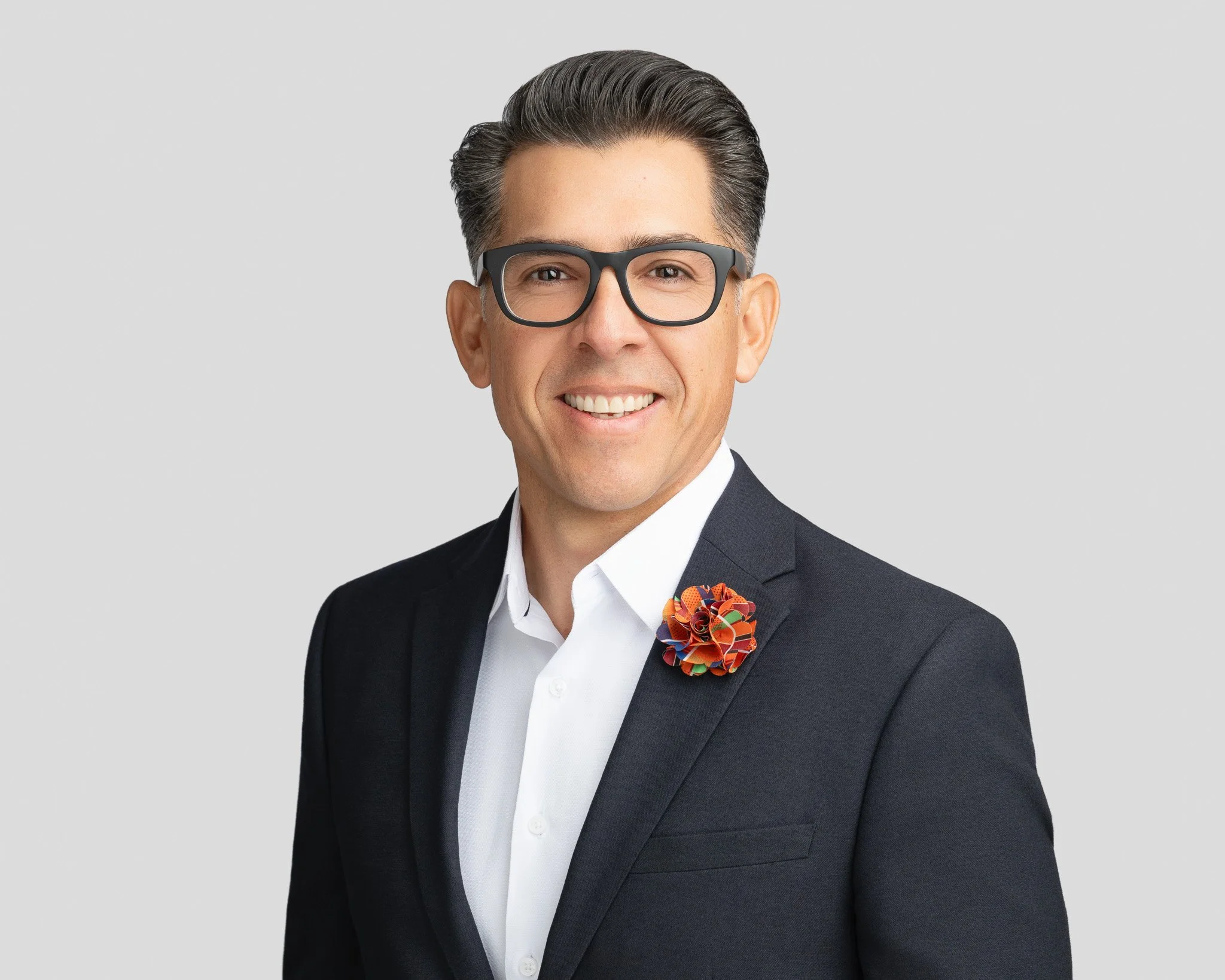 A smiling man with dark hair, glasses, wearing a dark suit and white shirt with a colorful brooch on his lapel against a plain gray background.