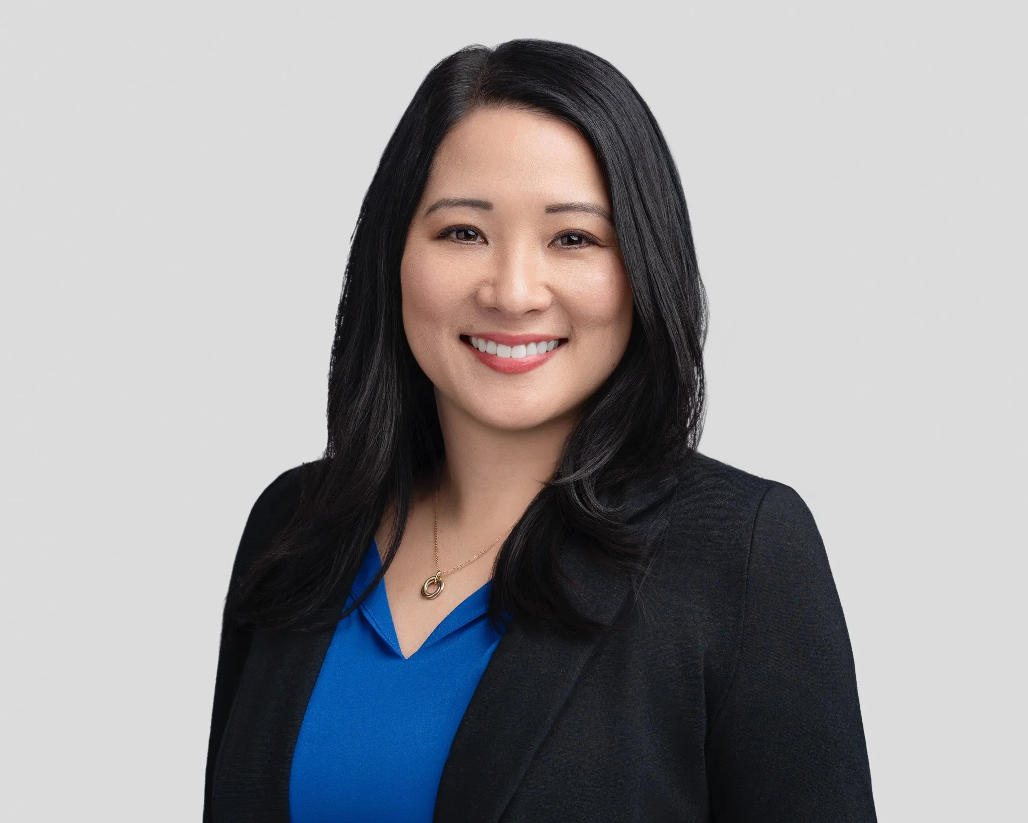 A professional woman with long black hair smiling, wearing a black blazer over a blue blouse with a gold necklace, against a light gray background.