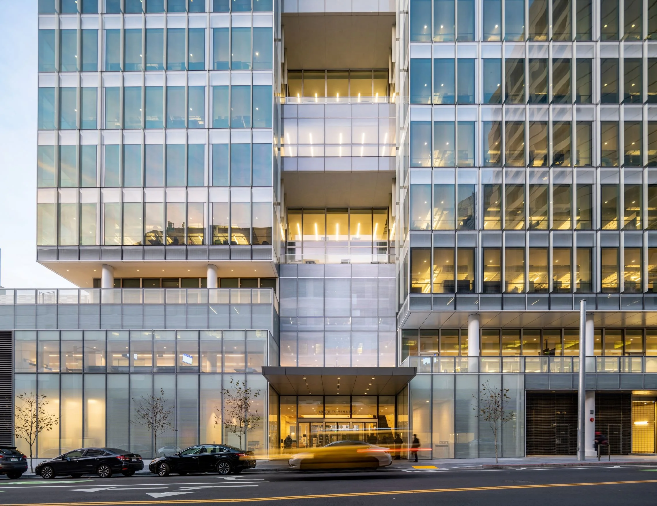 Modern multi-story office building with glass windows and illuminated interior, street view with parked cars and a moving yellow taxi in front.