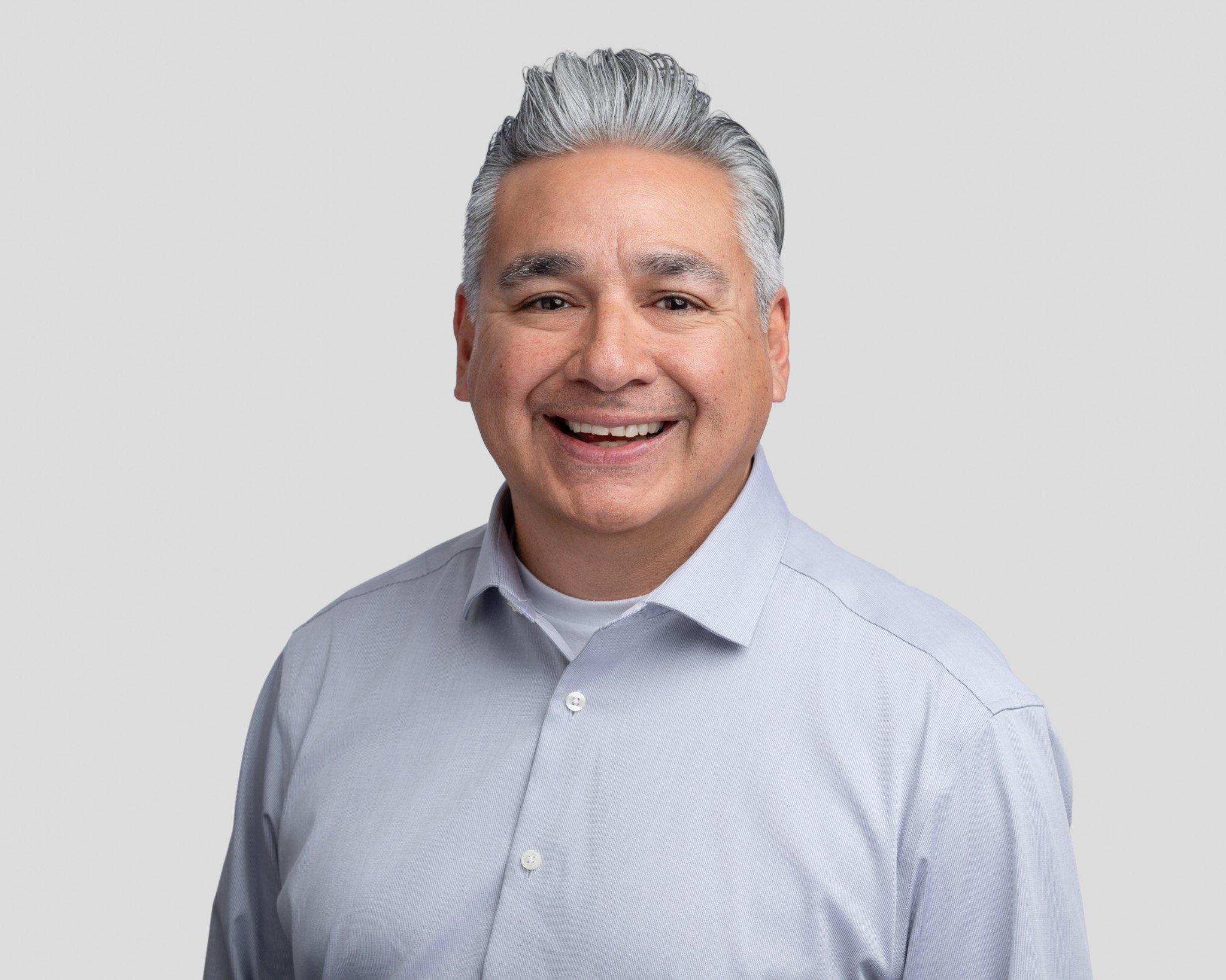Portrait of a smiling middle-aged man with gray hair, wearing a light blue collared shirt against a plain gray background.