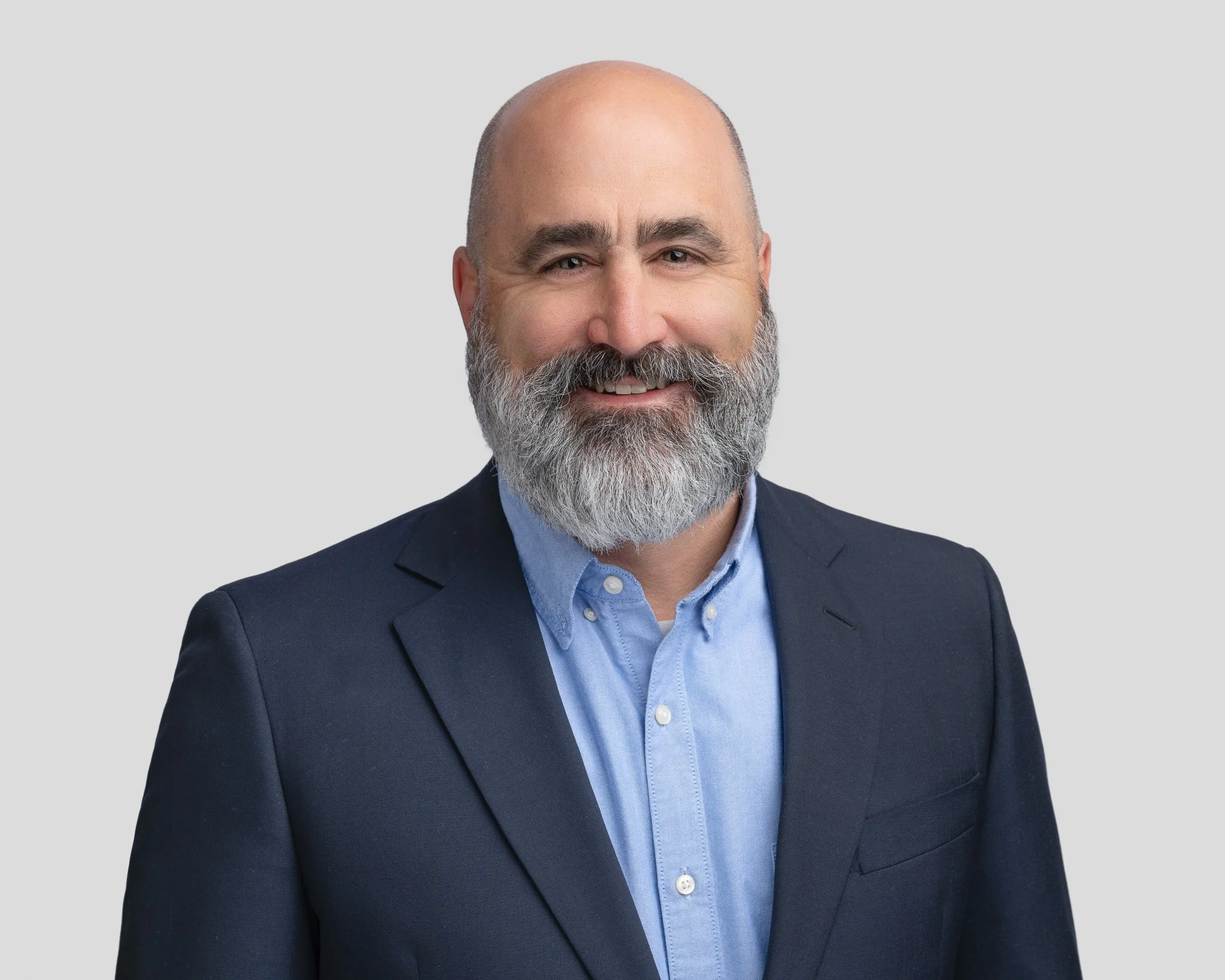 A middle-aged man with a beard and short hair in a business suit smiling at the camera against a plain light gray background.