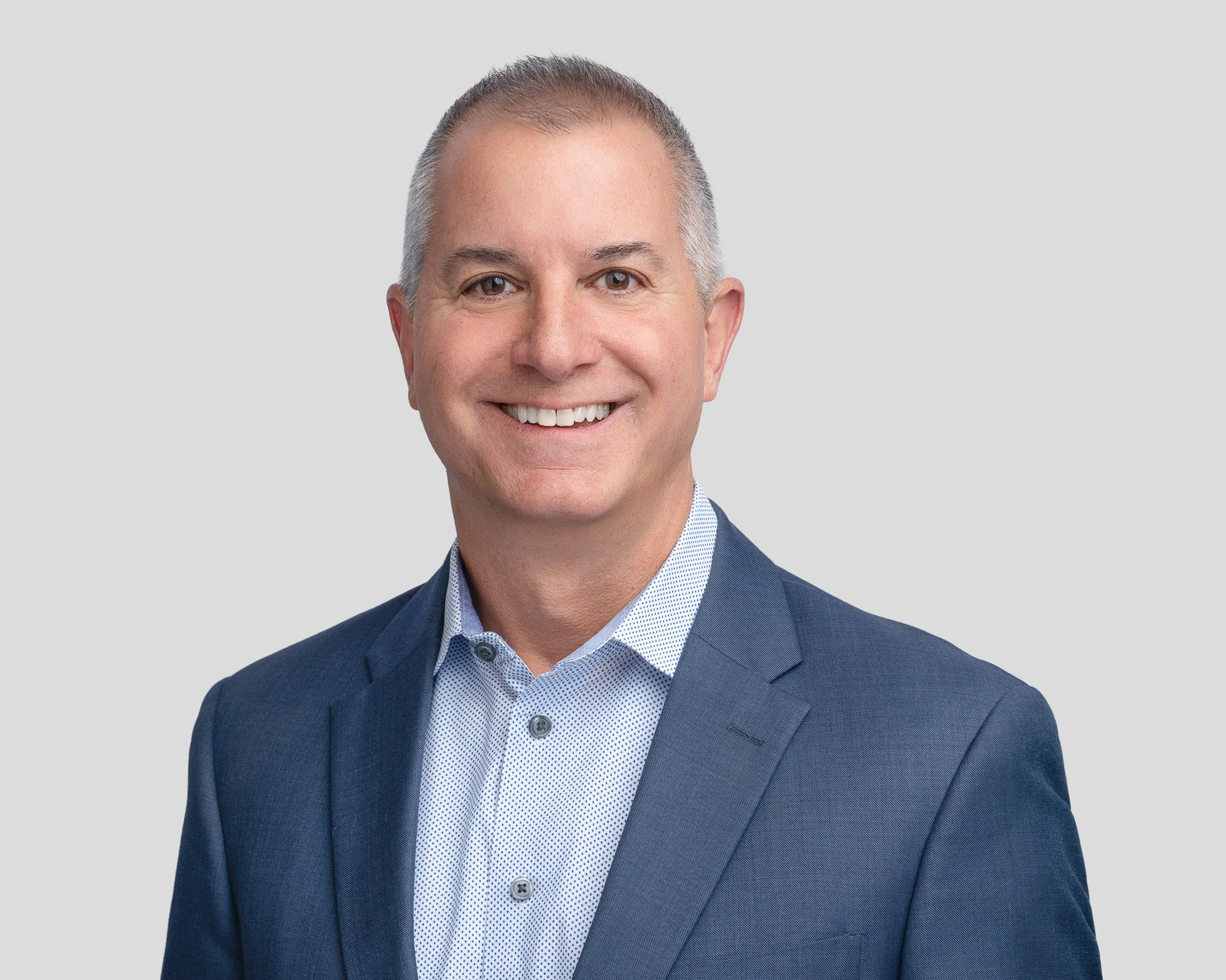 Headshot of a smiling man in a blue suit and light blue shirt against a plain background.