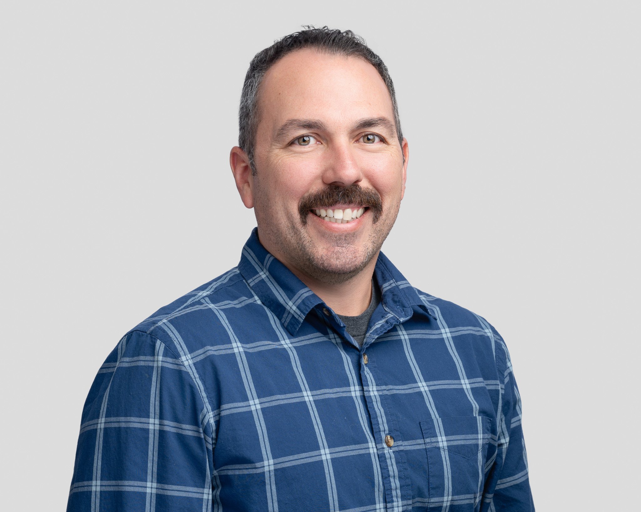 A man with short dark hair, facial hair, and a mustache, smiling, wearing a blue plaid shirt, against a plain light gray background.