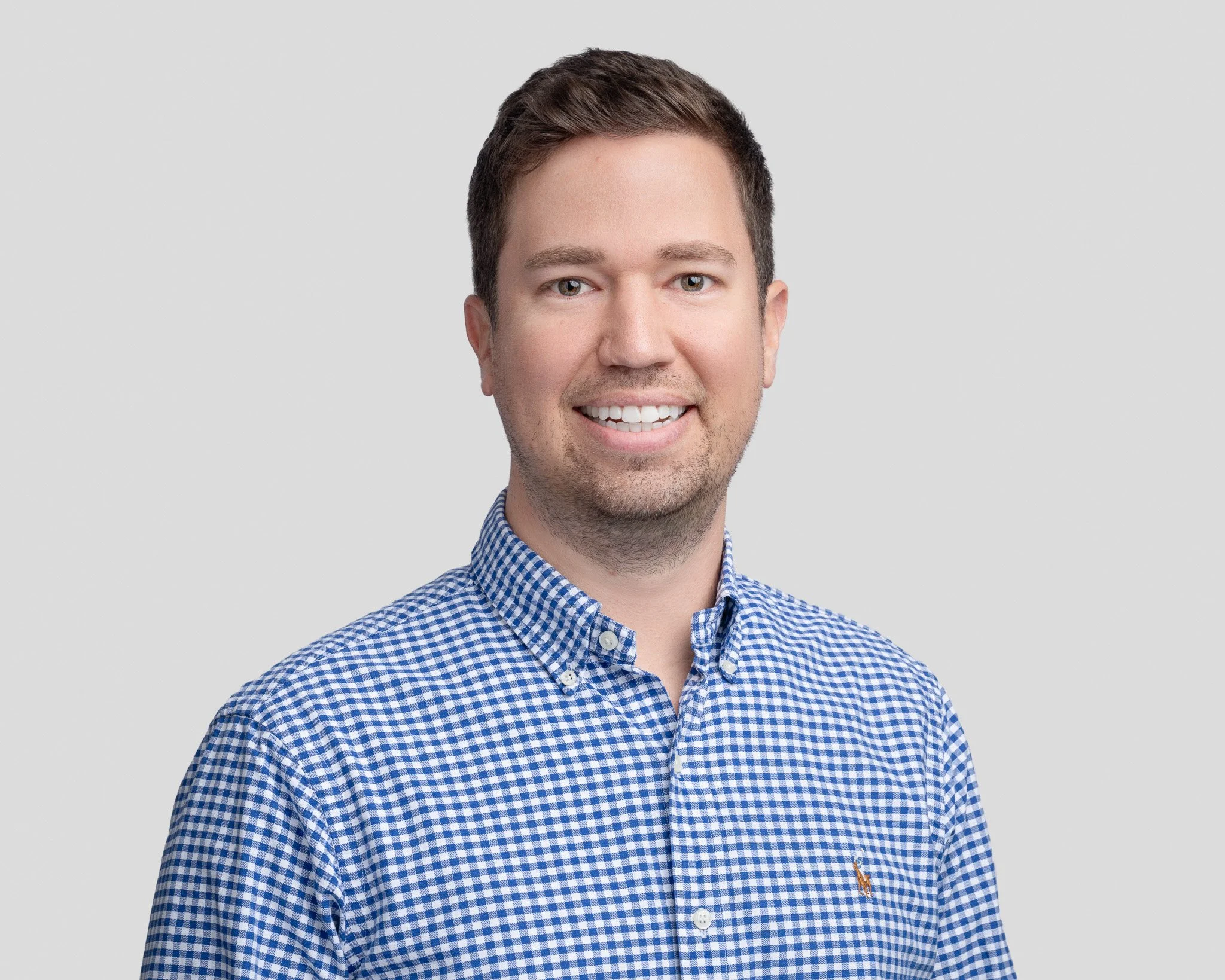 Portrait of a young man with short brown hair, smiling, wearing a blue and white checkered button-down shirt, against a plain light gray background.