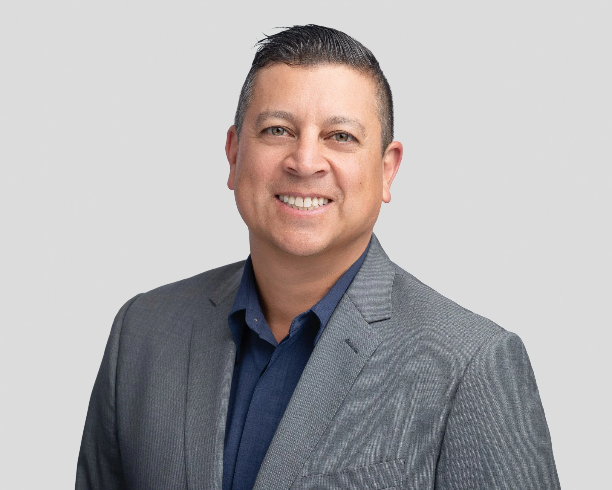 A professional man with short dark hair, wearing a grey suit and a navy blue shirt, smiling at the camera against a light grey background.
