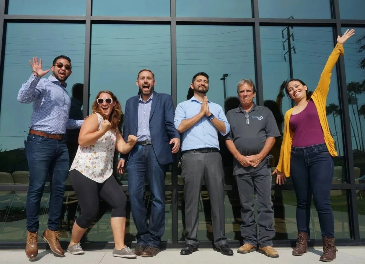 Six diverse people standing outside in front of a glass building, smiling and celebrating, some with arms raised and others with hands clasped or fists clenched.