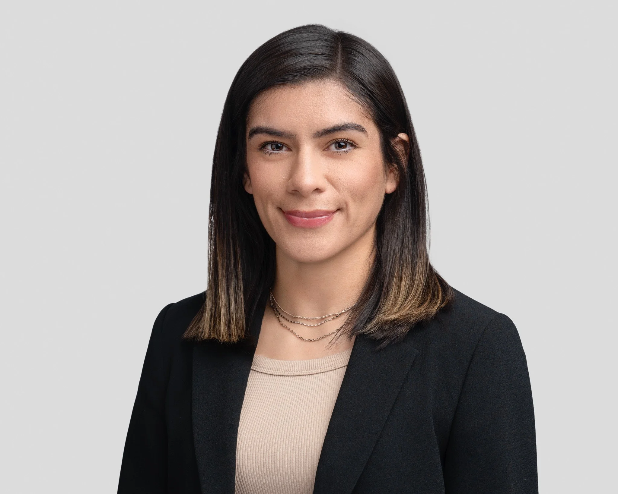 A professional young woman with straight dark hair, wearing a black blazer and beige top, smiling against a plain light gray background.