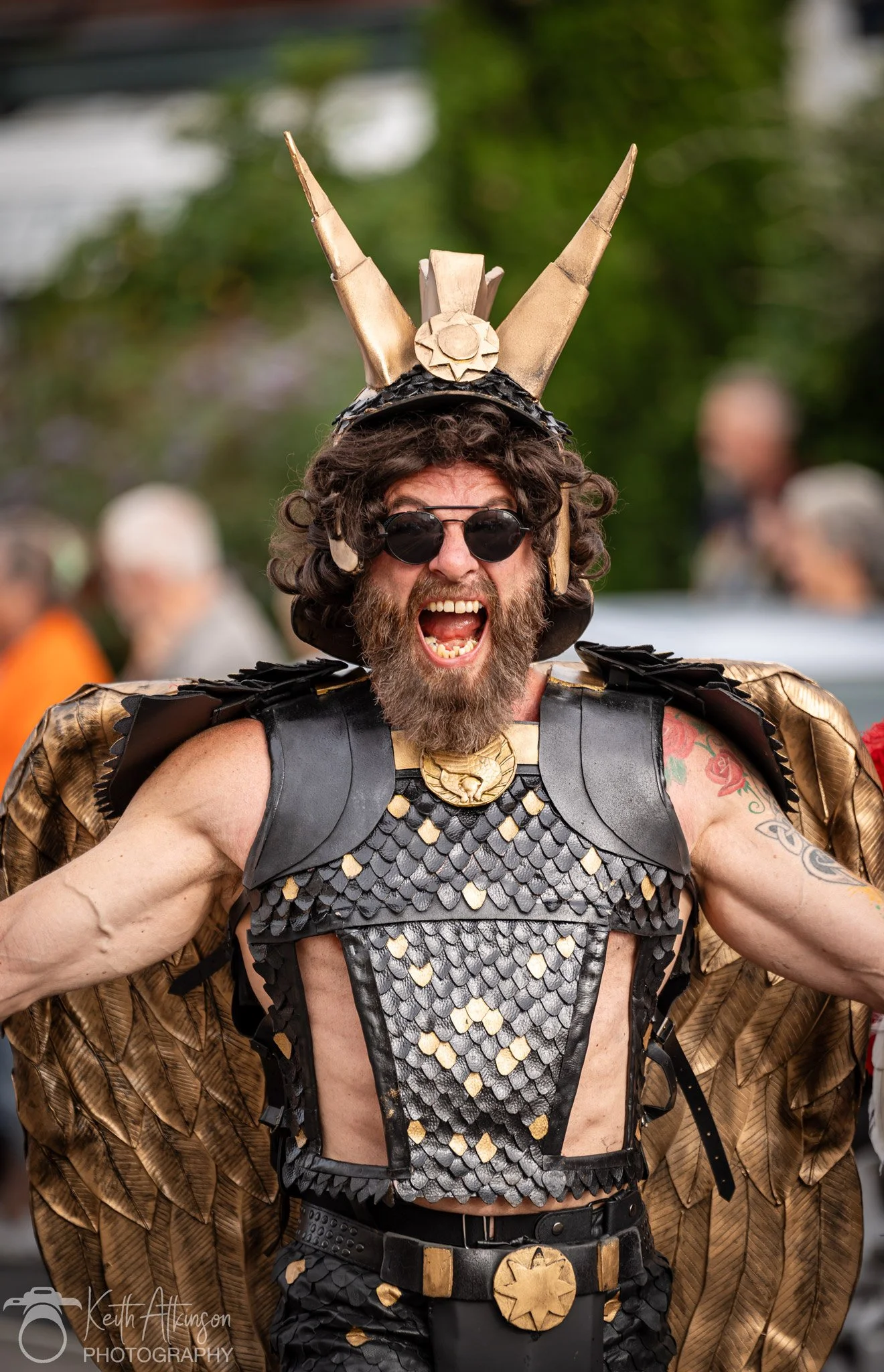 A man wearing a costume with a gold and black scale pattern, gold shoulder details, and a large gold headpiece with horns. He has curly hair, a beard, sunglasses, and is shouting with his mouth open.