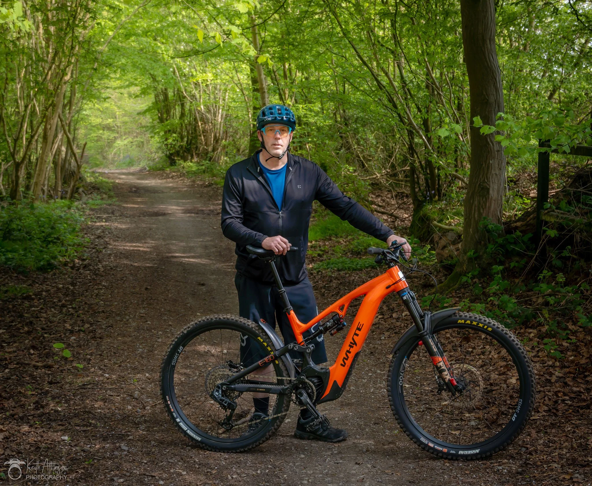Man in black jacket and blue helmet standing next to an orange mountain bike on a dirt trail in a lush green forest.