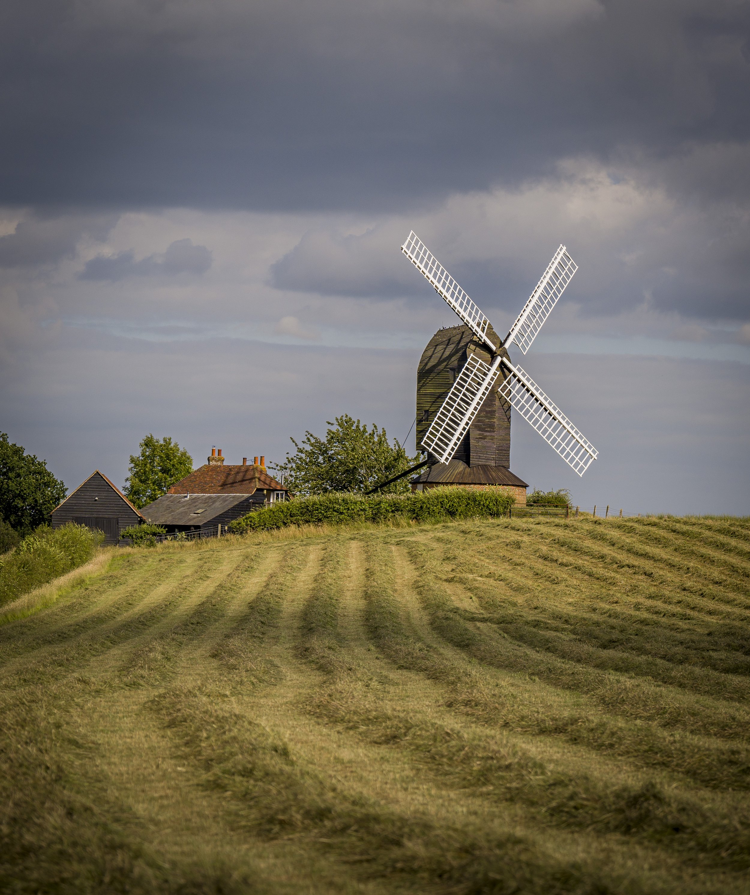 A traditional wooden windmill on a hilltop under stormy clouds, surrounded by a fenced pasture and nearby houses with tiled roofs.