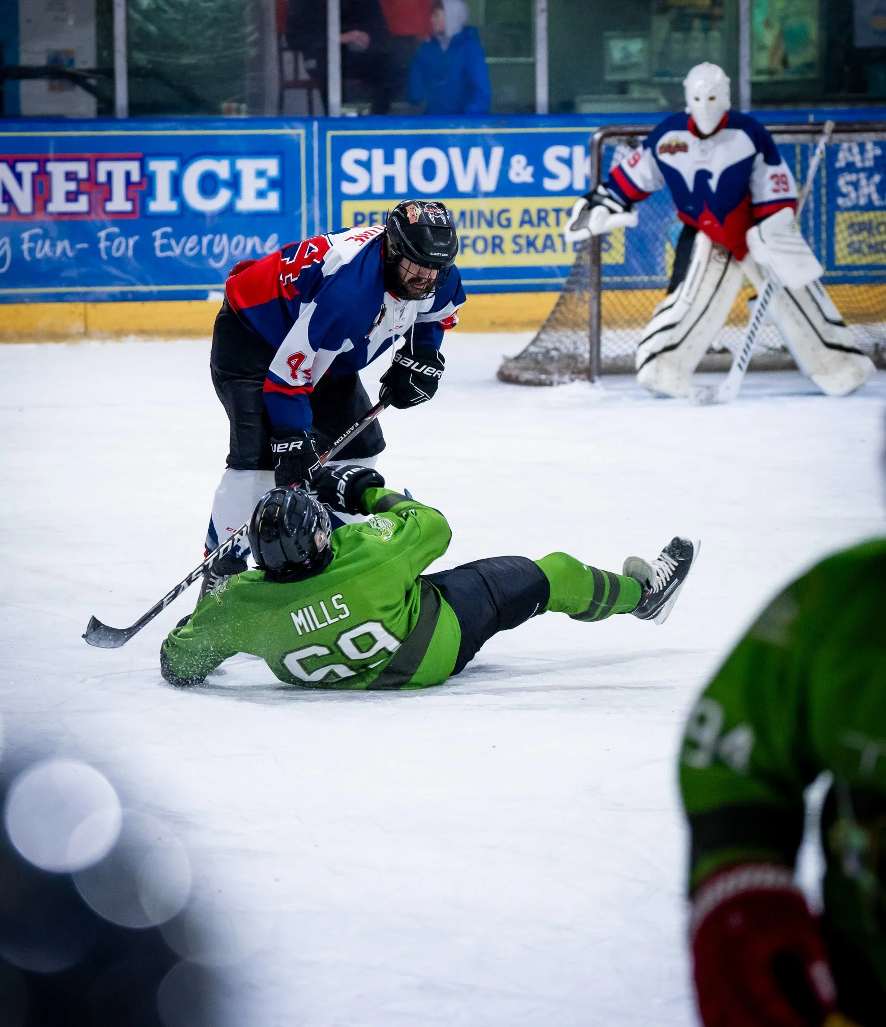 An ice hockey game action shot showing a player in a green jersey on the ice, lying on the rink with the name 'Mills' and number 60, being challenged by an opponent in a blue, red, and white jersey. The opponent is standing and leaning over, enacting