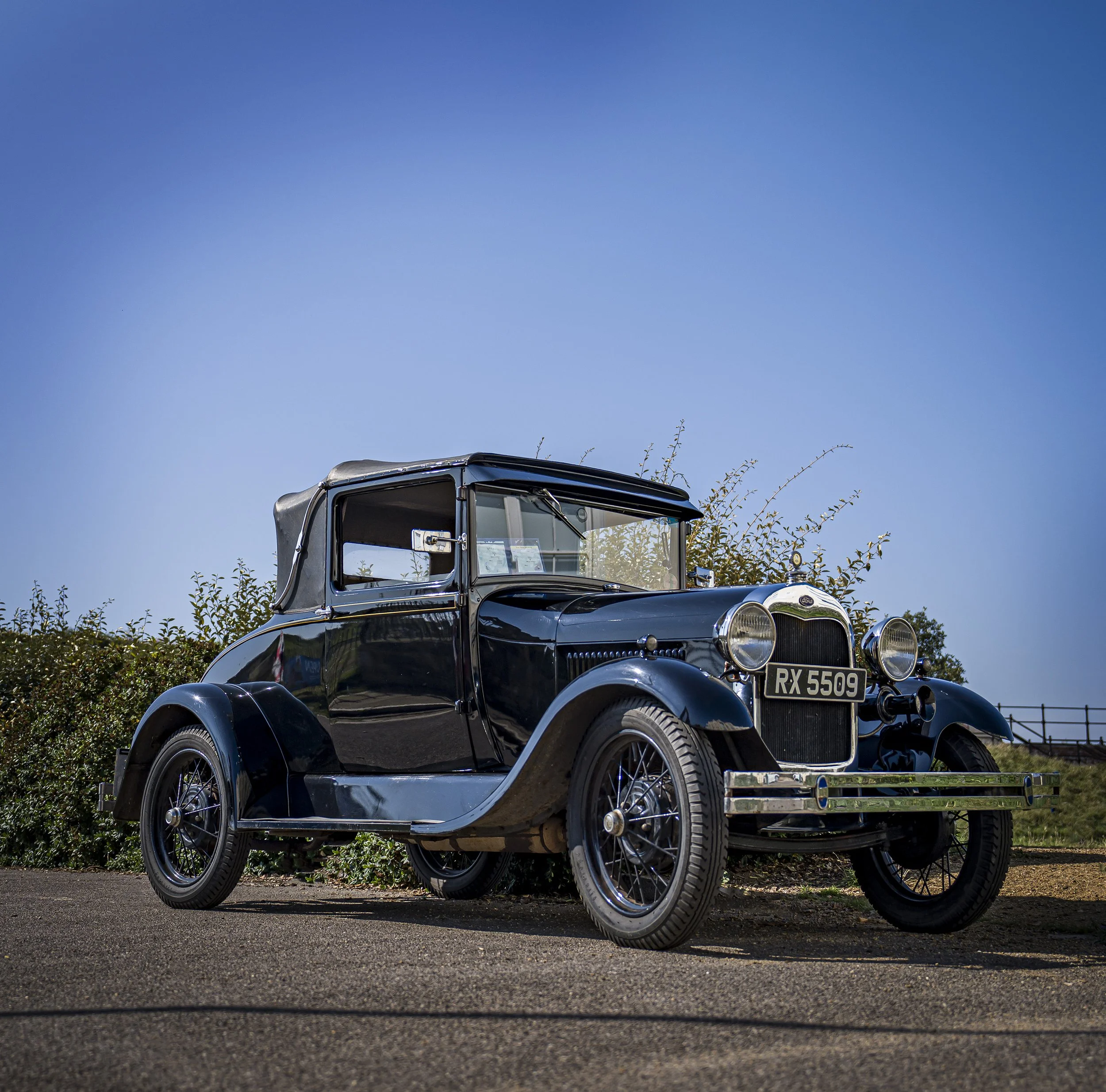 A vintage black car with a soft top roof parked on the side of a road under a clear blue sky.