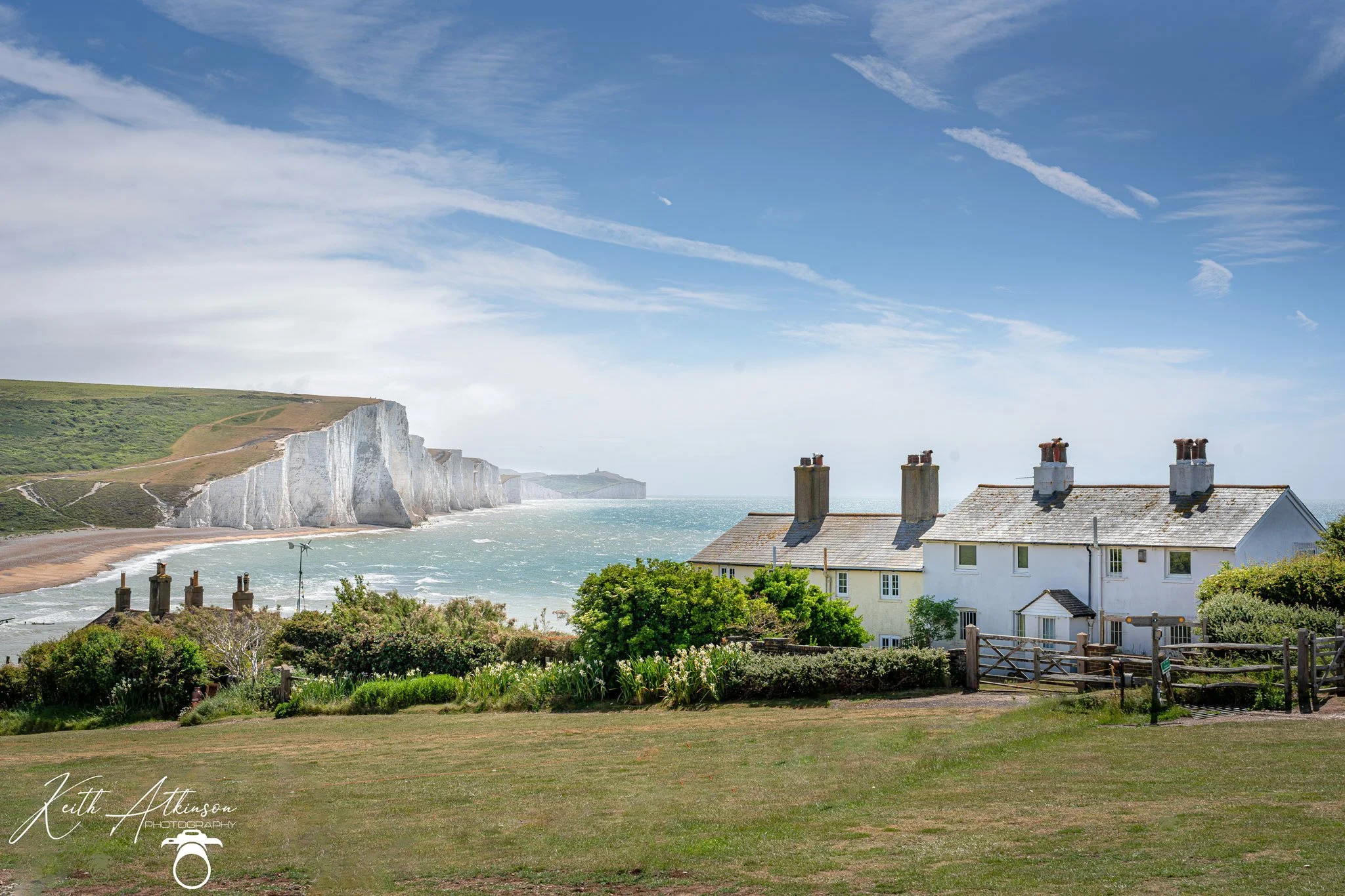 Coastal view of the seven sisiters white chalk cliffs, ocean, and farmland, with a row of houses with chimneys in the foreground under a partly cloudy sky.