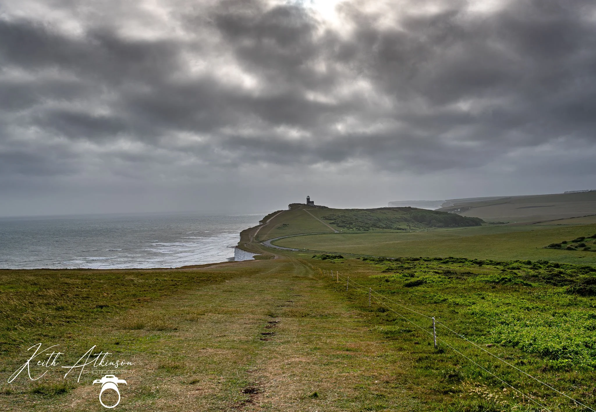 A scenic east sussex coastal landscape with lush green fields, a winding path, and the Belle Tout lighthouse on a hill under a dark, cloudy sky.