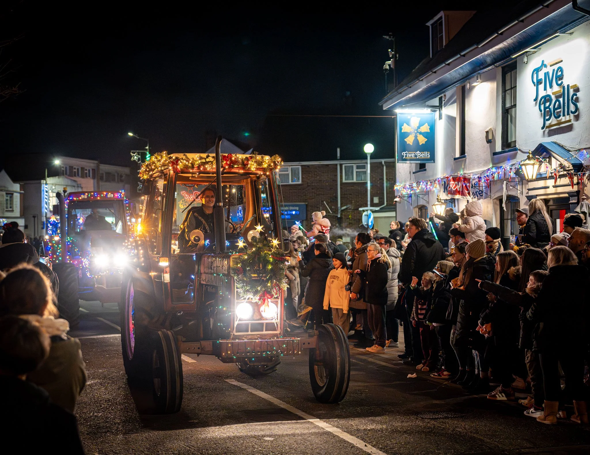 A nighttime parade with decorated tractors, a crowd of spectators, and a building with a sign that says "Five Bells" decorated with Christmas lights.
