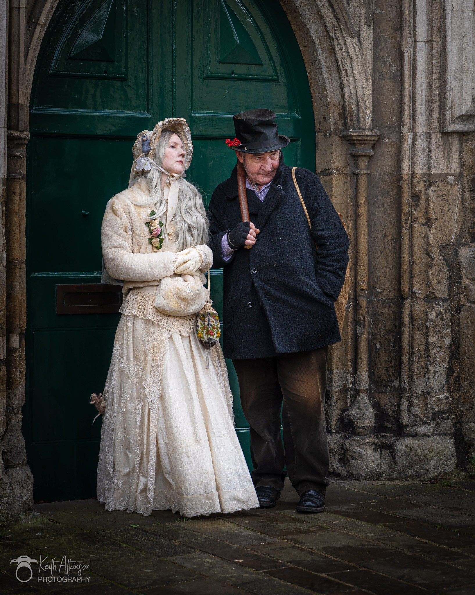 Two people dressed in historical clothing standing near a green door on a stone building. One person is wearing a vintage cream dress, gloves, and a bonnet; the other is wearing a black coat and hat, holding an umbrella.