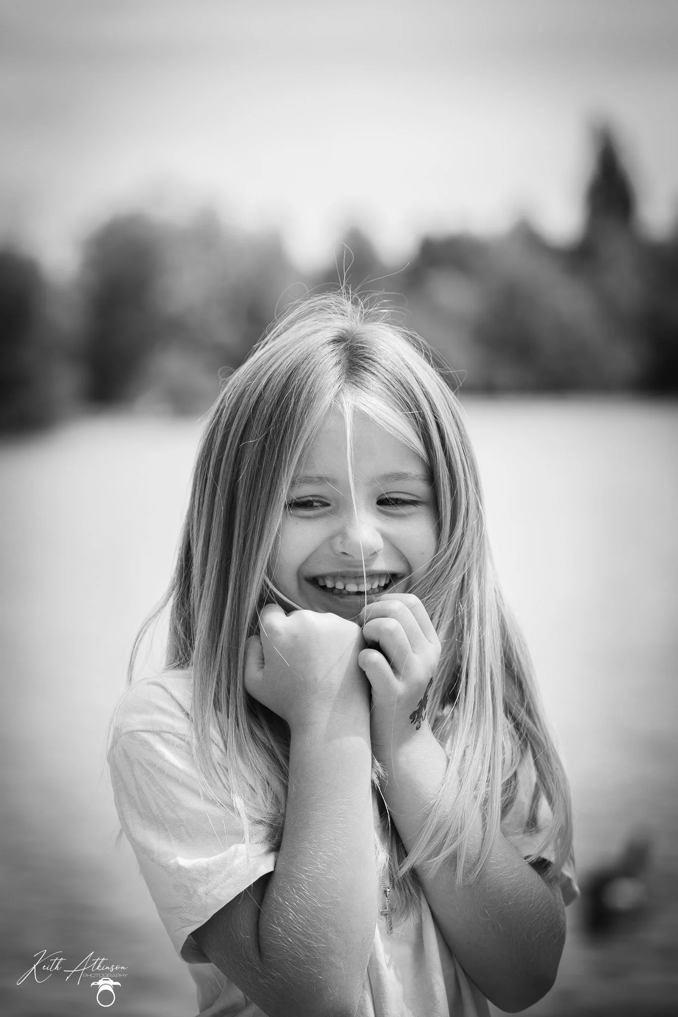 A smiling young girl with long hair playing outdoors, holding a string or wire near her face, with a blurred background of trees and sky in black and white.