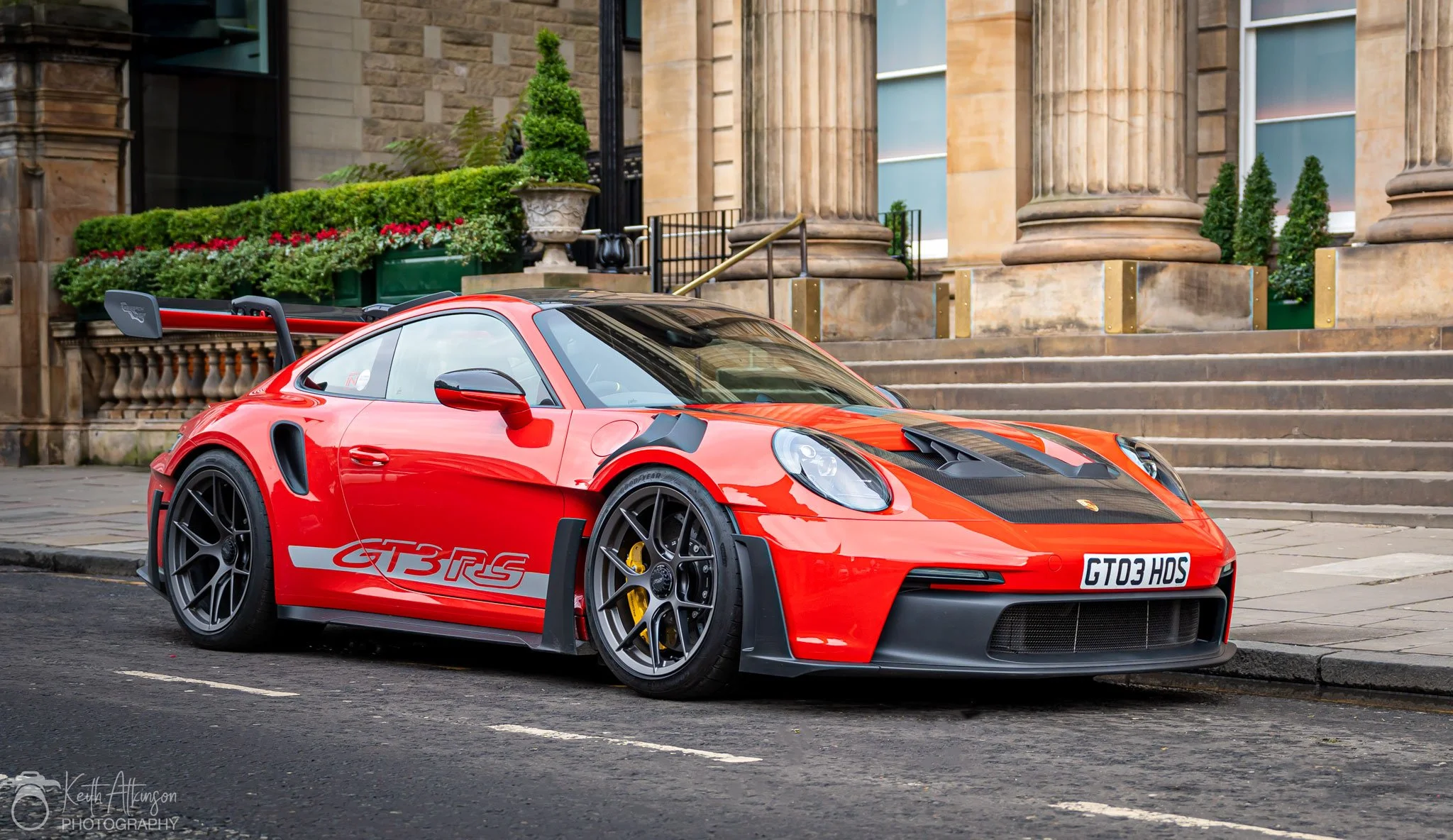 A red Porsche 911 GT3 RSR sports car parked on a city street in front of historic buildings with columns, stairs, and potted plants.