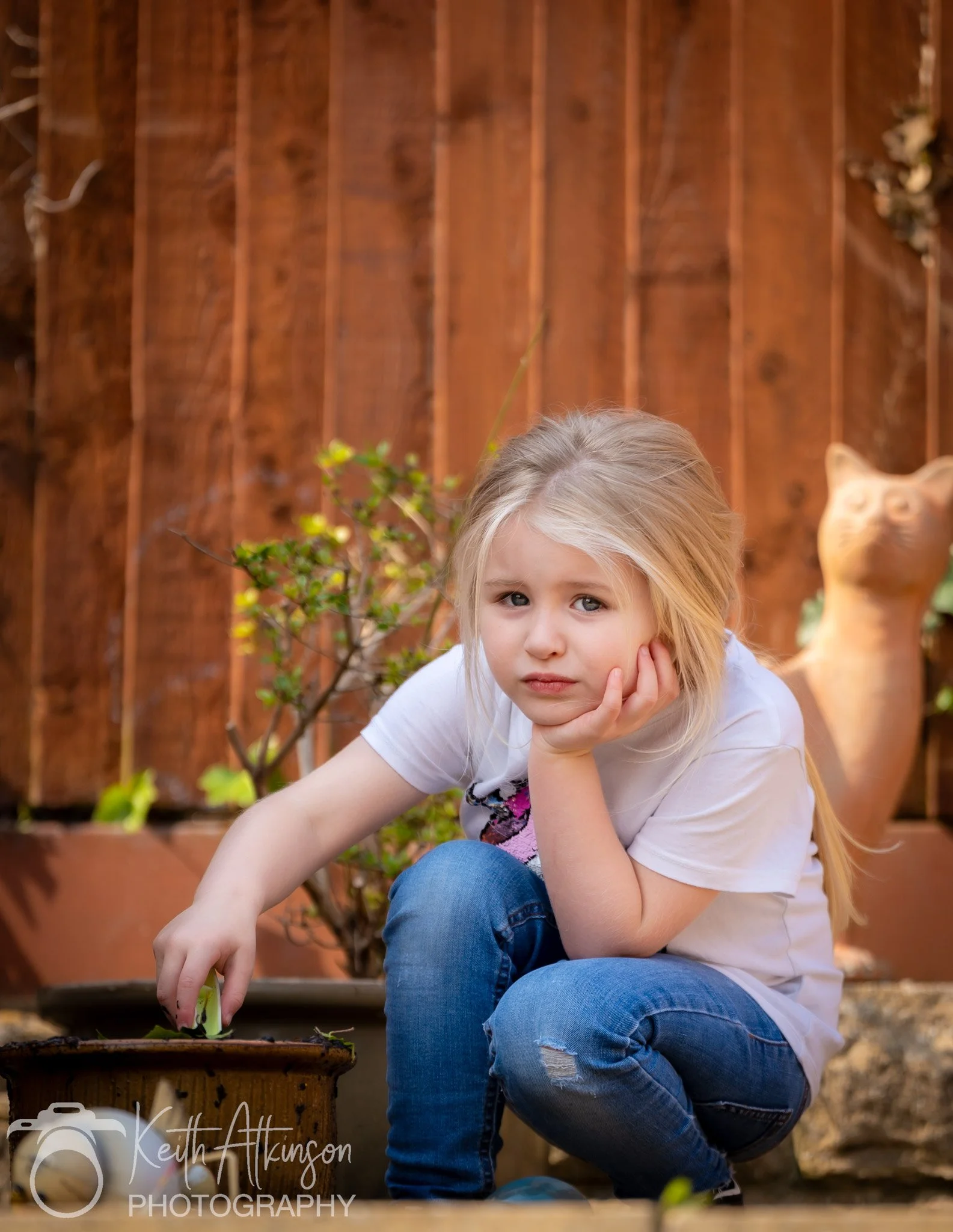 A young girl with long blonde hair, wearing a white T-shirt and ripped blue jeans, sits outdoors next to a wooden fence. She has a bored or upset expression, resting her face on her hand, with her other hand touching a plant in a pot.