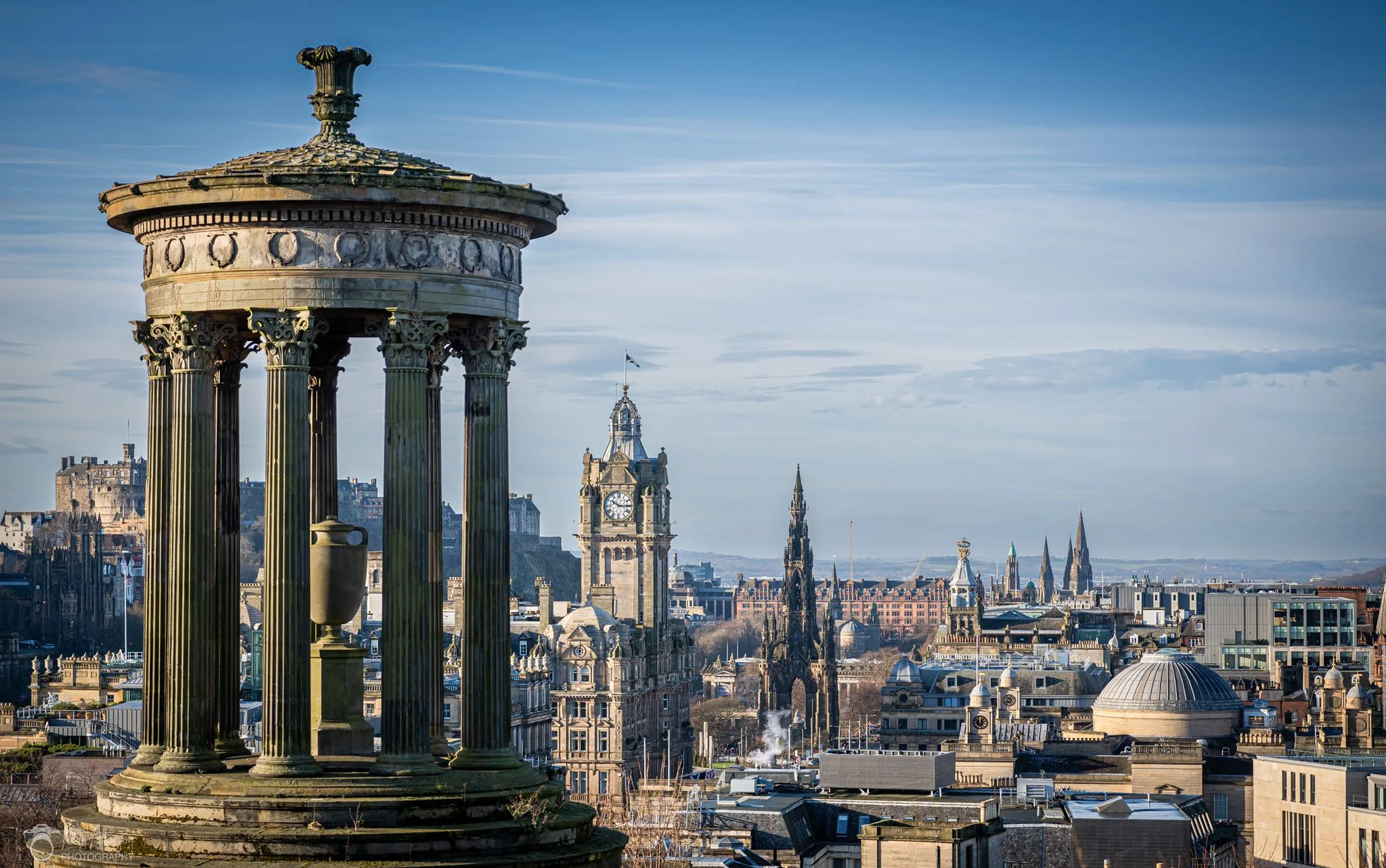 View of Edinburgh city skyline with the Dugald Stewart Monument in the foreground, showing classical columns and a decorative top, and historic buildings and spires in the background under a clear blue sky.