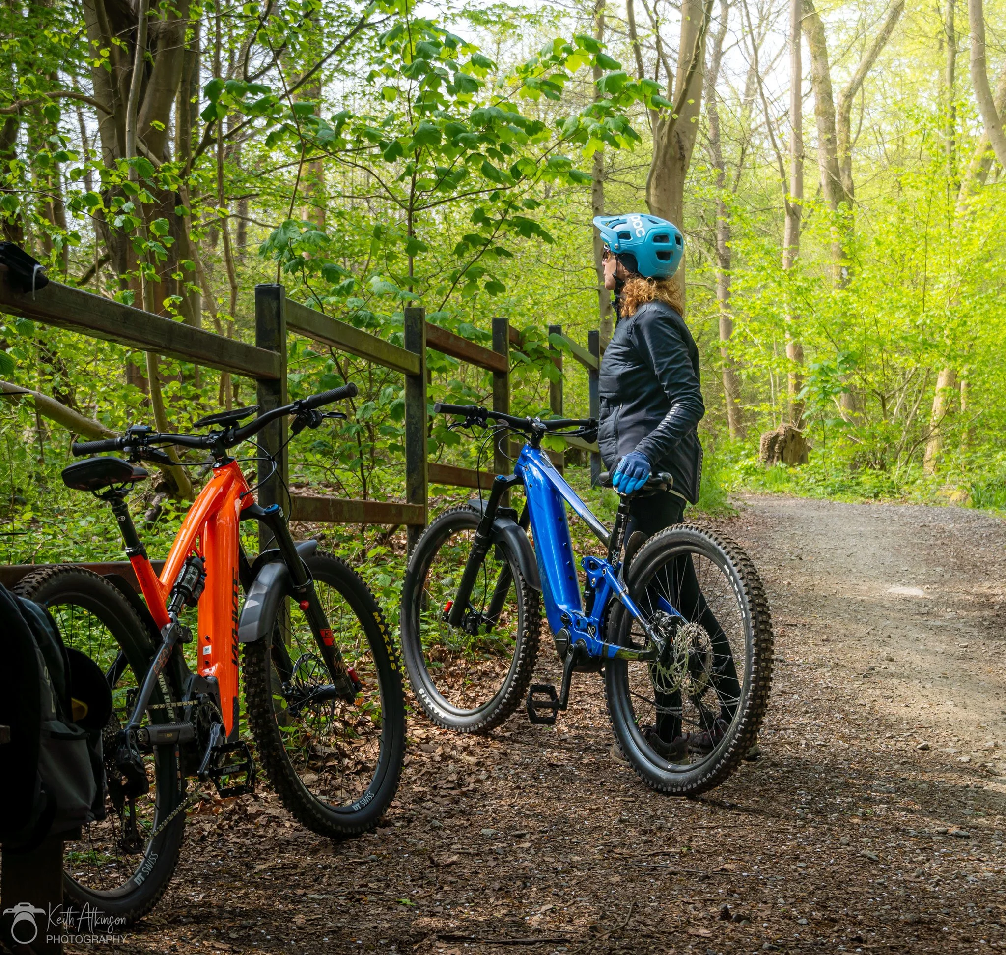 A woman wearing a blue helmet and black jacket standing with her blue mountain bike on a forest trail, with another orange mountain bike nearby.