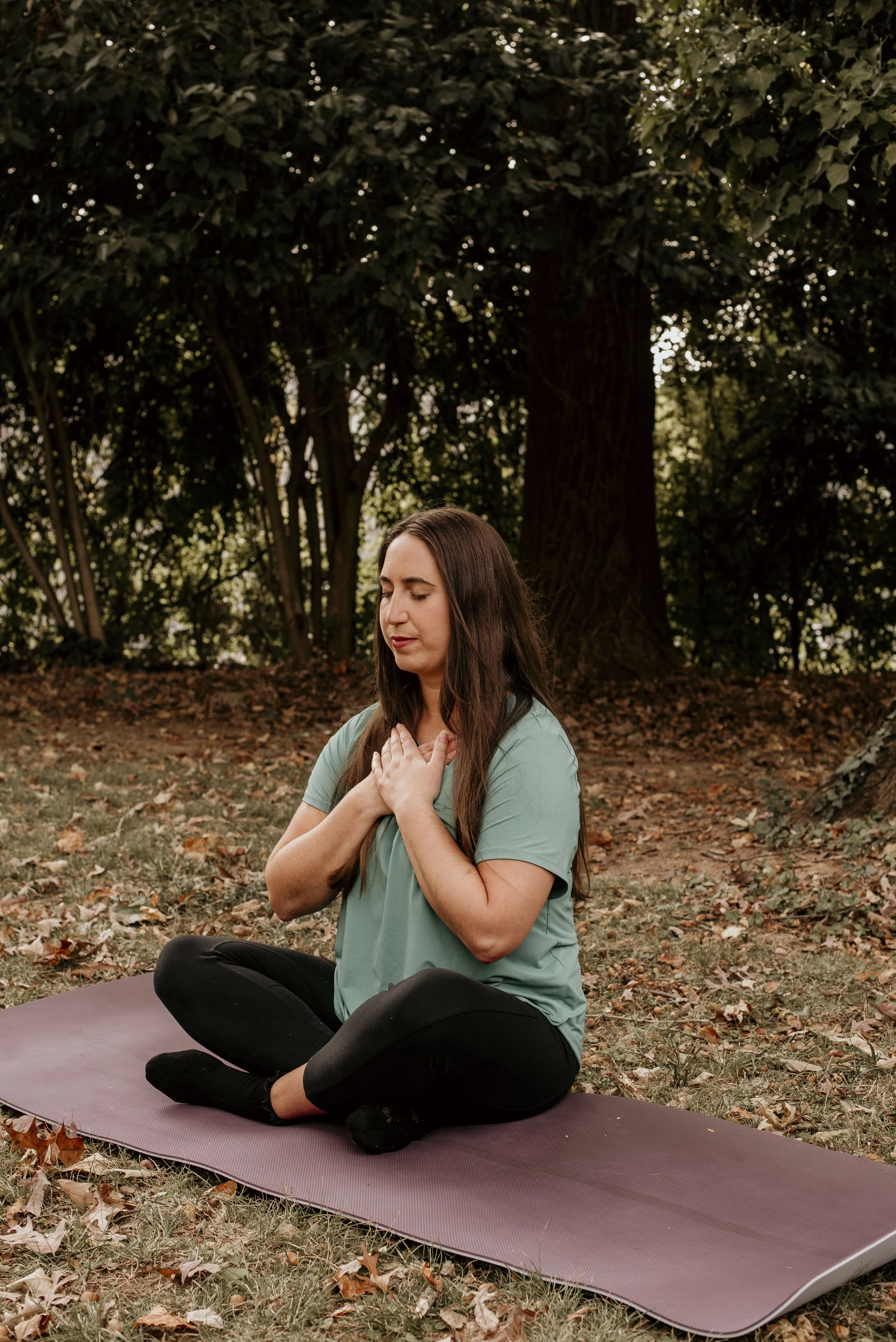 A woman practicing yoga outdoors on a mat, sitting cross-legged with hands on her chest, eyes closed, surrounded by trees and fallen leaves.
