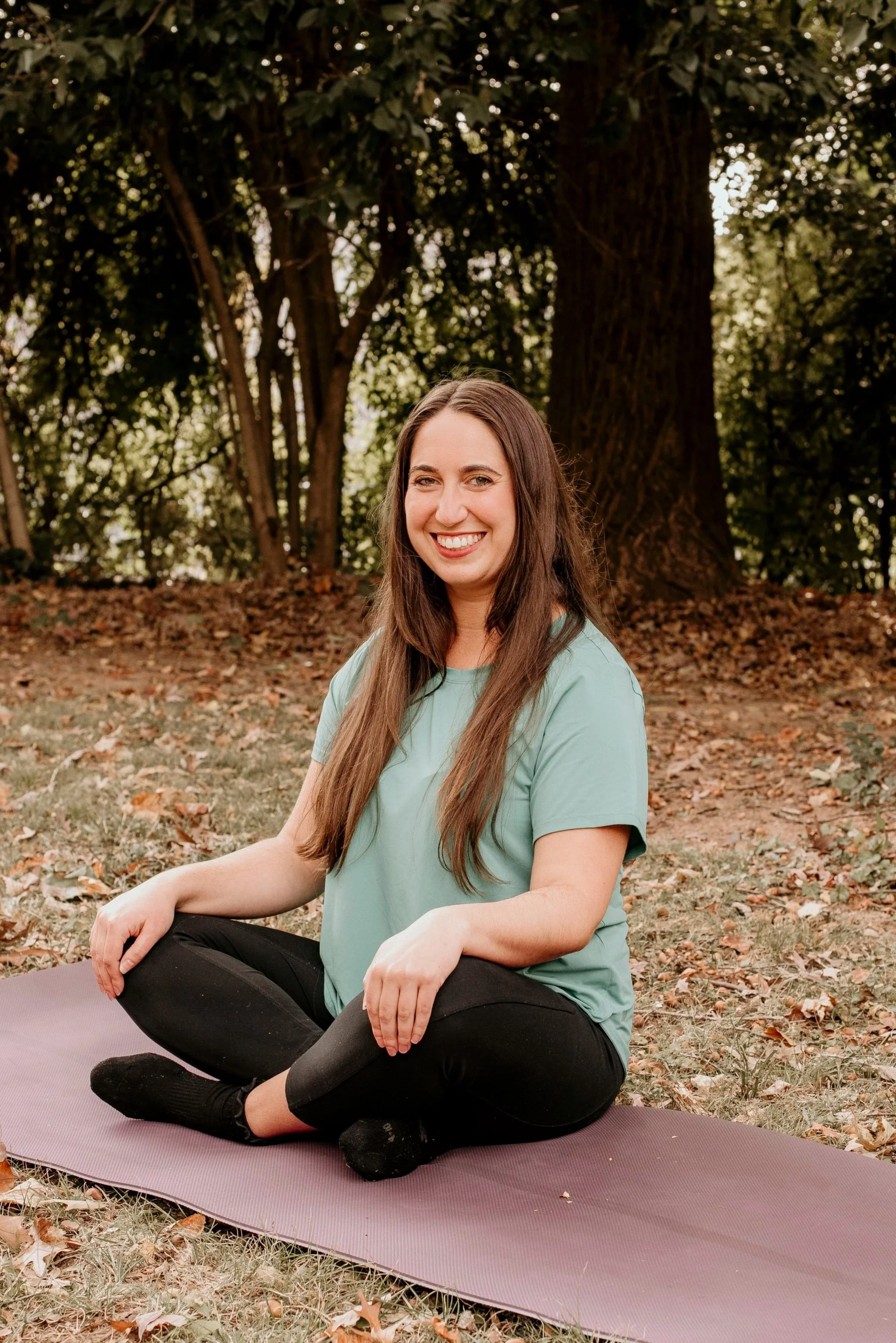 A woman practicing yoga outdoors on a purple yoga mat, sitting cross-legged on grass with fallen leaves, smiling at the camera with trees in the background.