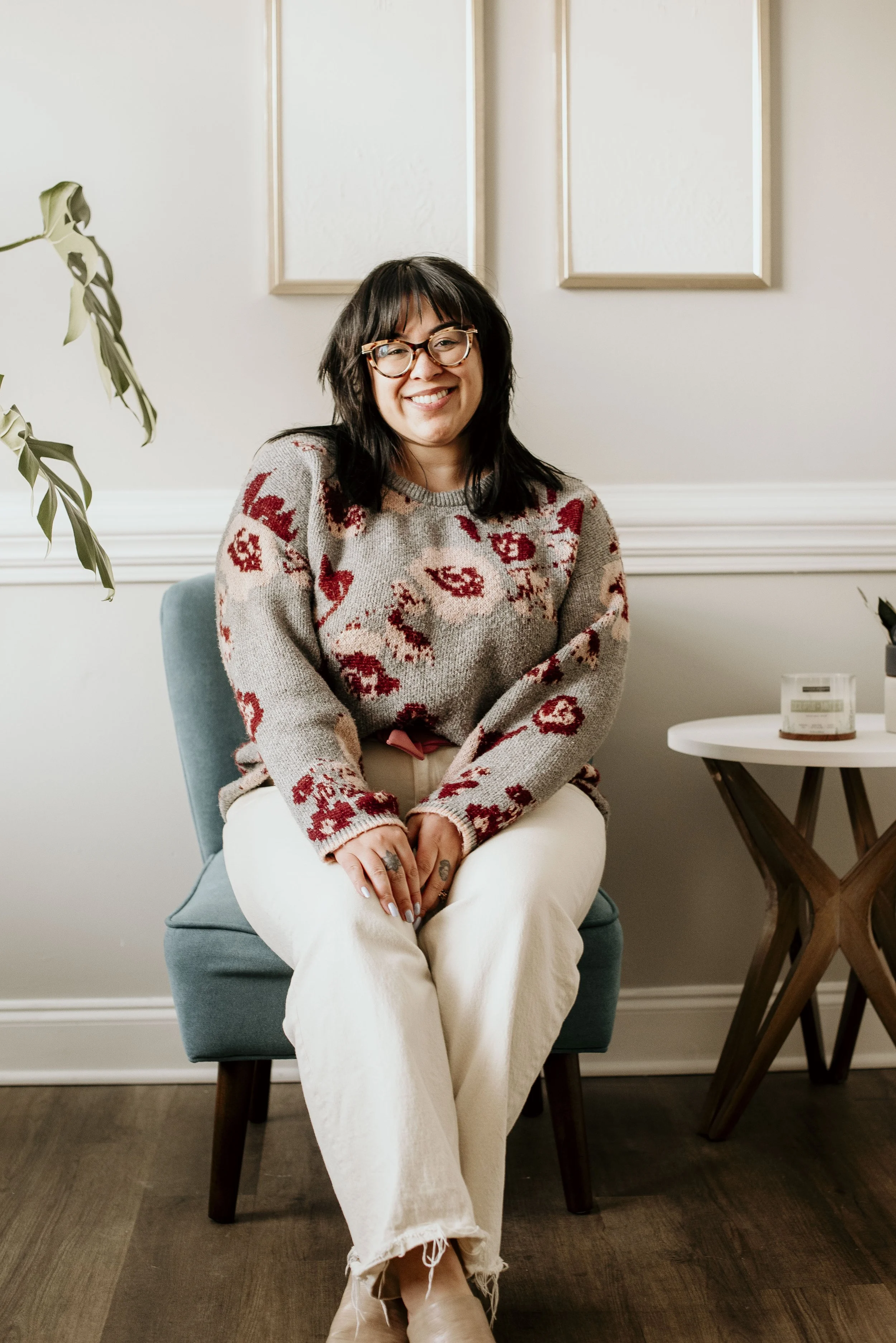 A woman with glasses and dark hair, smiling, sitting on a blue chair in a room with beige walls and framed artwork. She is wearing a patterned sweater and cream-colored pants.