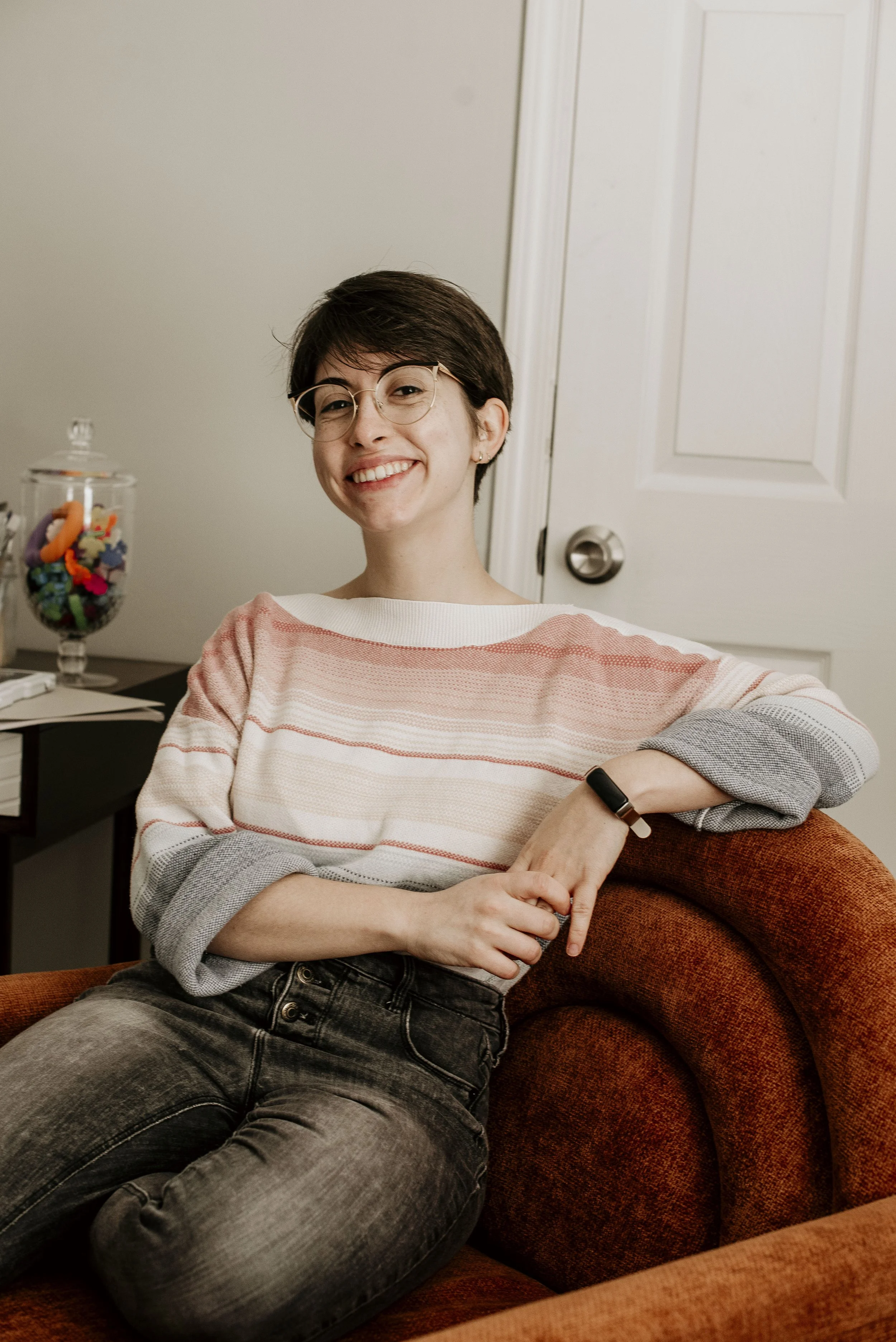 A young woman with short brown hair, glasses, and a striped sweater, smiling and sitting on a rust-colored armchair in a cozy room, with a white door behind her and a jar of colorful candies on the side table.