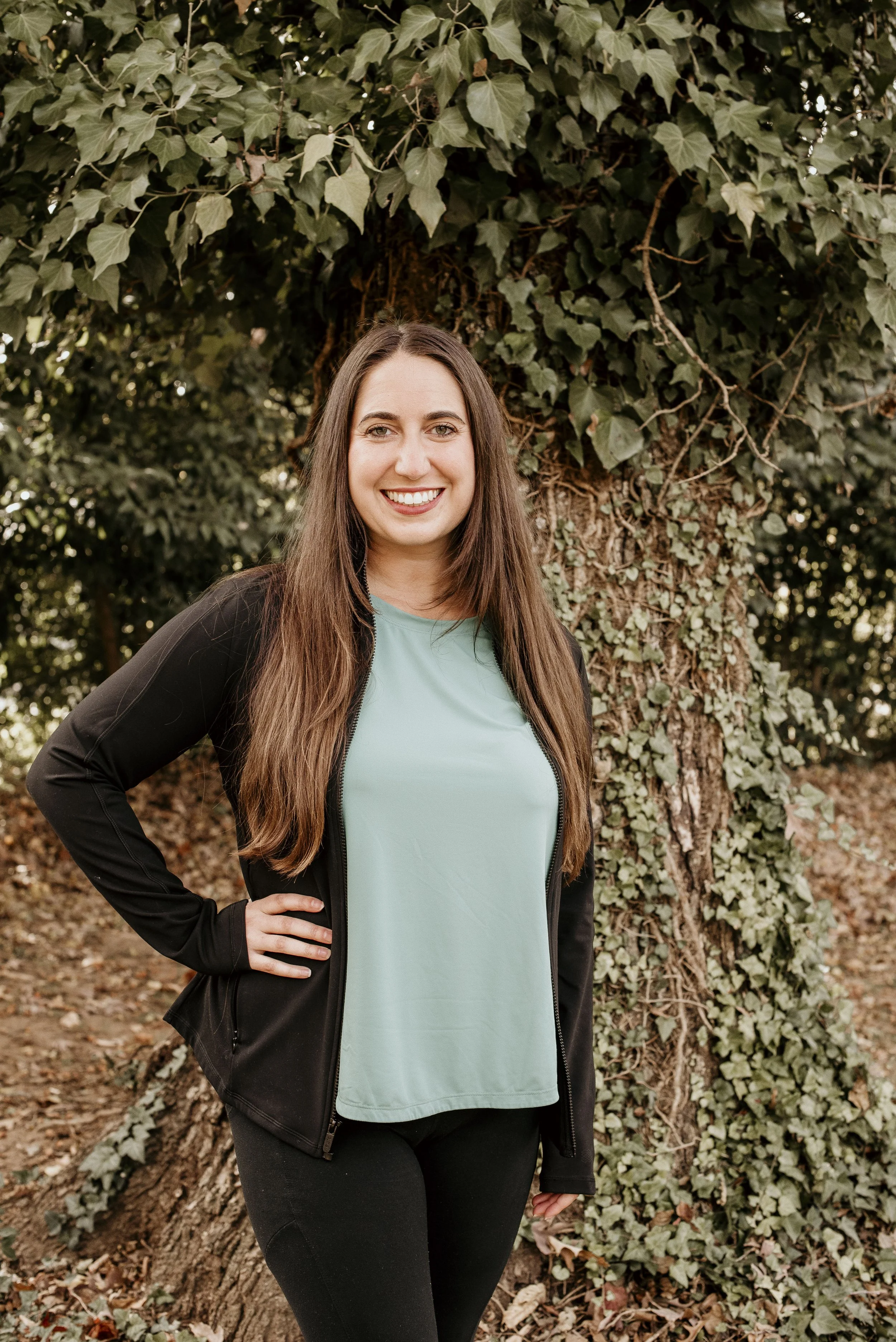 A young woman with long brown hair smiling, wearing a black jacket over a light green shirt, standing outdoors in front of a large ivy-covered tree.