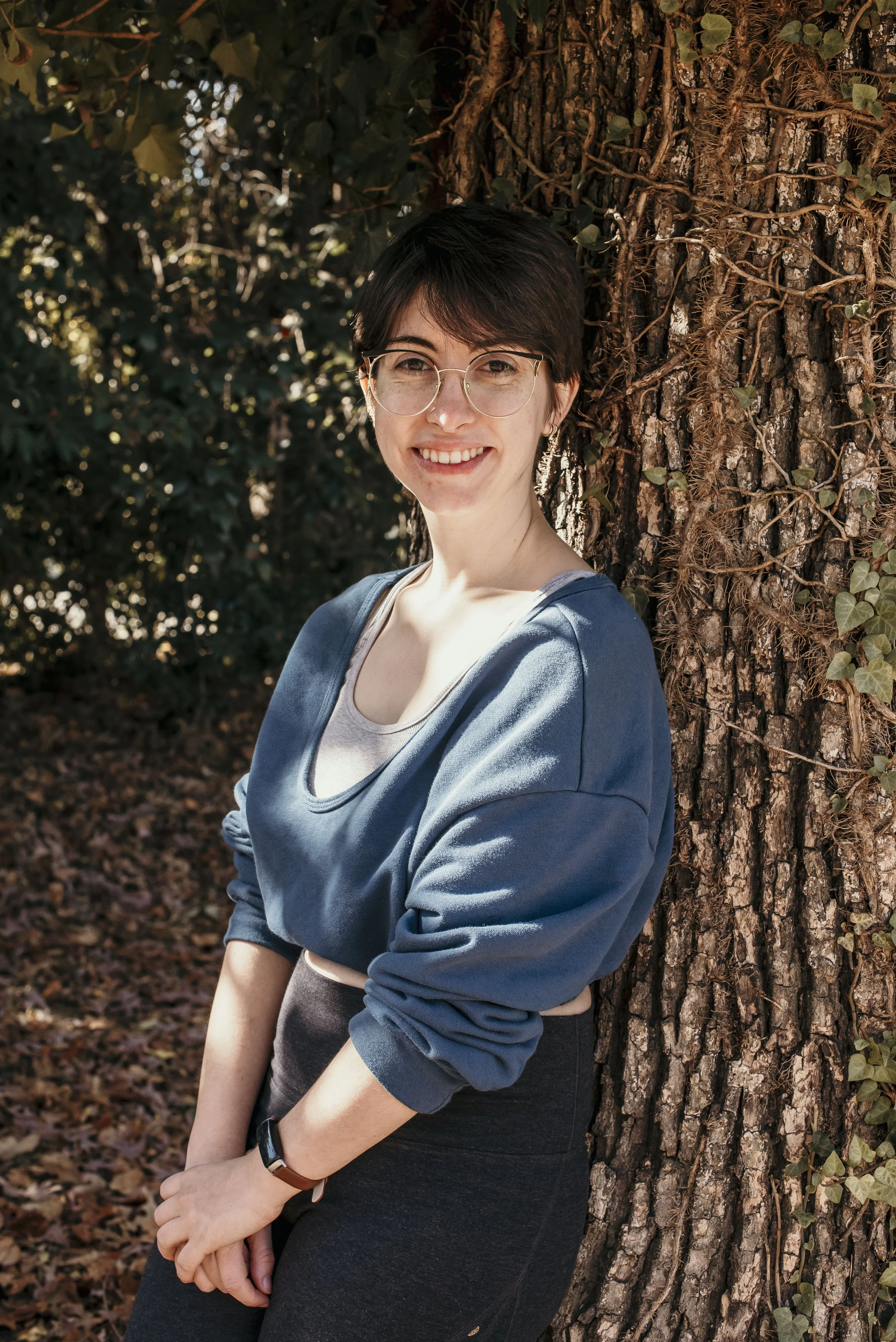 A young woman with short dark hair and glasses leaning against a tree outdoors. She is smiling and wearing a blue sweatshirt over a light top, with a black wristwatch, surrounded by autumn leaves and greenery.