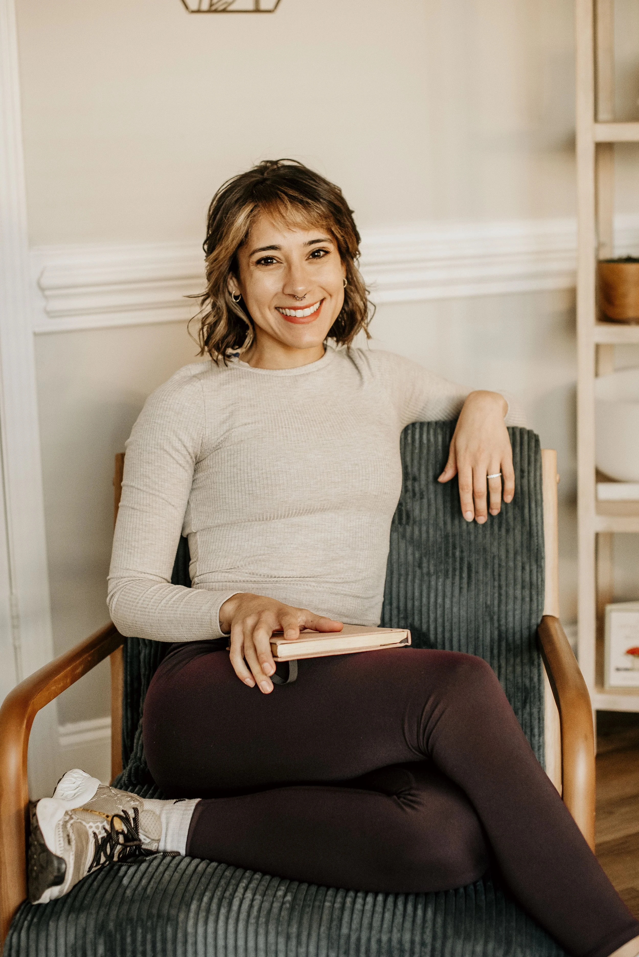 A young woman with dark wavy hair, glasses, and tattoos on her arms and torso, smiling and posing outdoors by a stone wall. She is wearing a light green crop top and blue shorts.