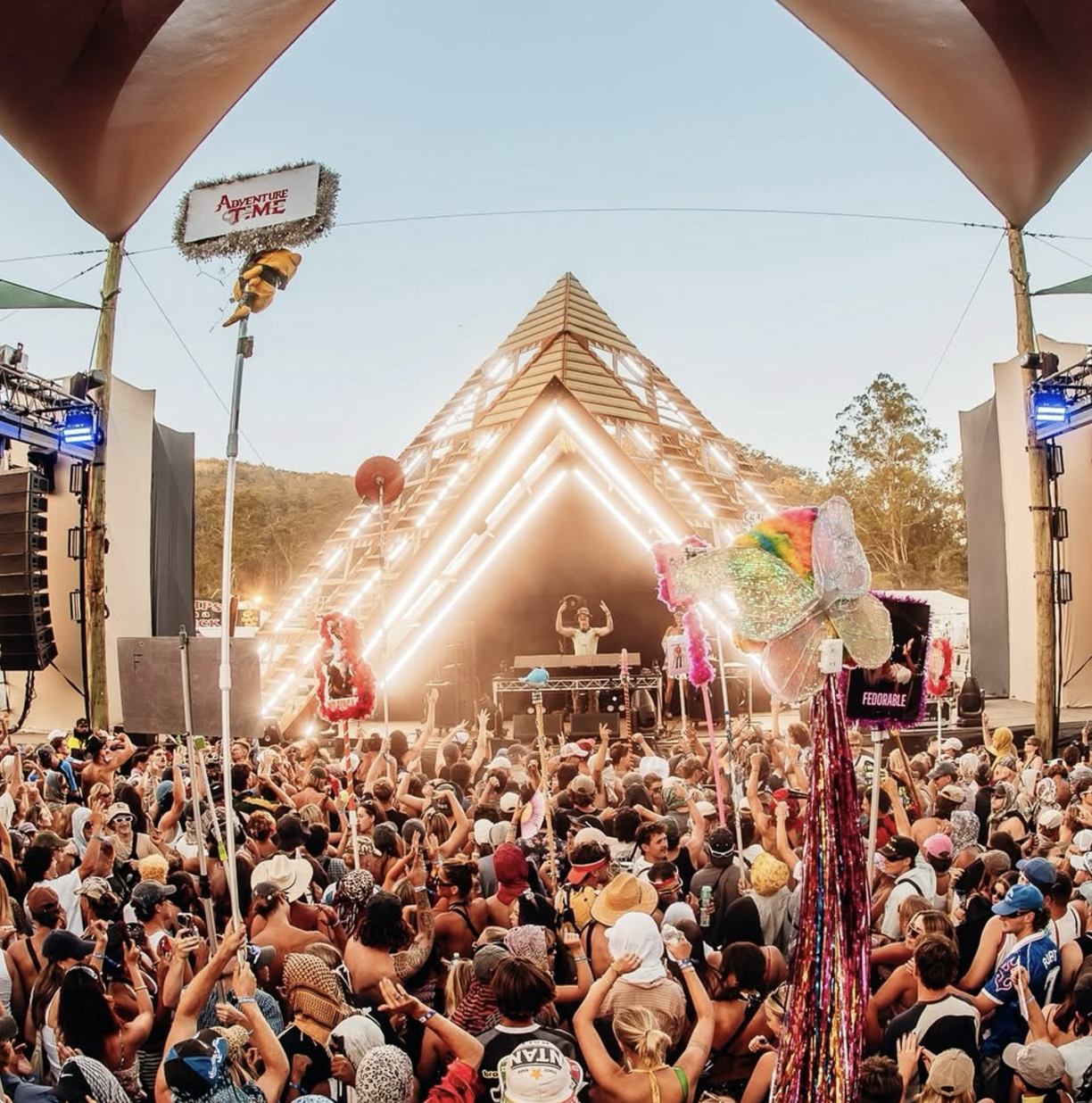 Crowd gathered at an outdoor music festival with a DJ on stage, illuminated pyramid-shaped stage structure, and colorful decorations including a large butterfly statue and feathers.