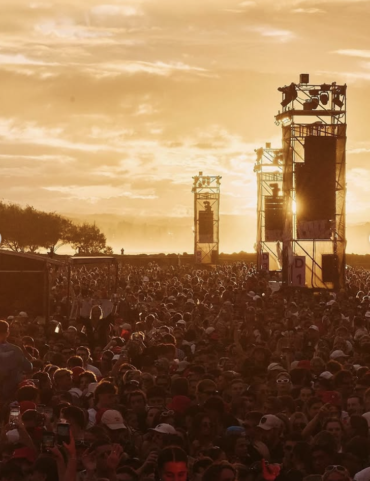 Crowd of festival attendees facing outdoor stage at sunset with scaffolding and large speakers.