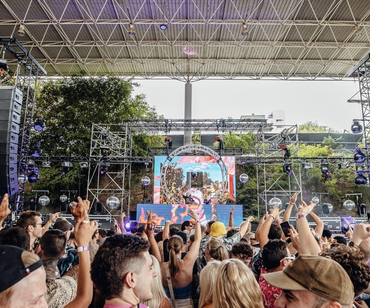 Crowd at an outdoor music concert with stage, LED screens, and dancing people.