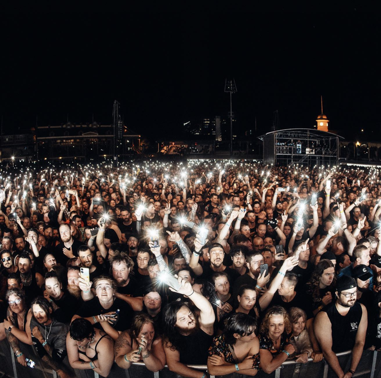 A large crowd of concert attendees at night, many holding up smartphones with flashlight features on in the air during a live music event.