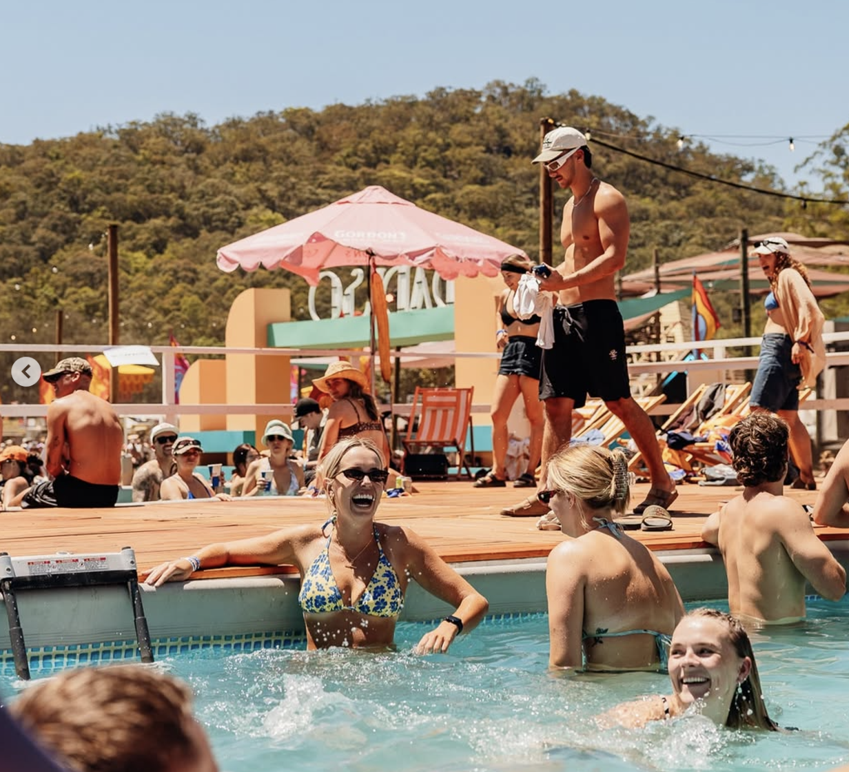 People enjoying a pool party on a sunny day, with some standing on the pool deck while others swim in the pool, surrounded by colorful umbrellas and lounge chairs, hills in the background.
