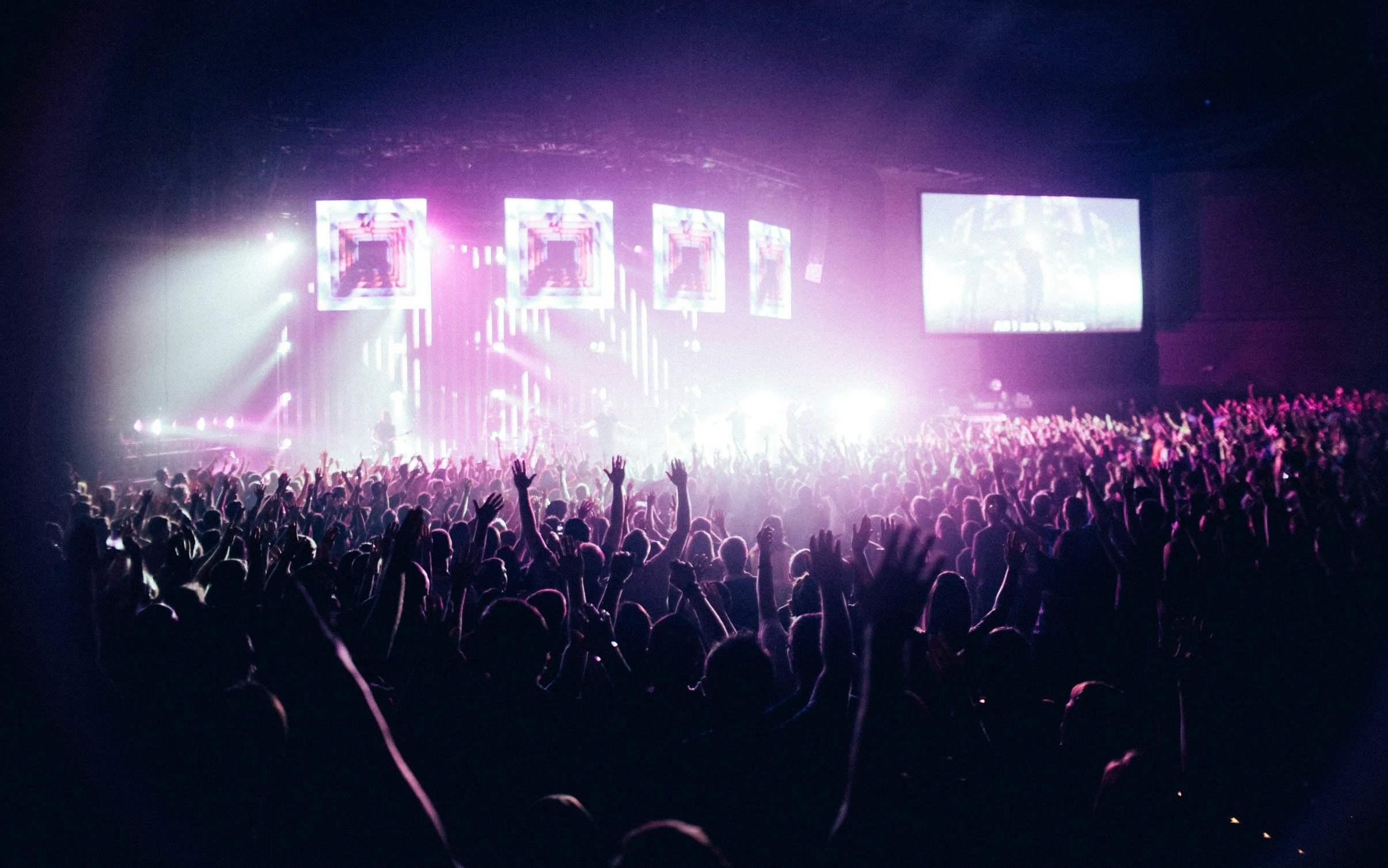 Crowd of people at a concert or music event with colorful stage lighting and large screens.