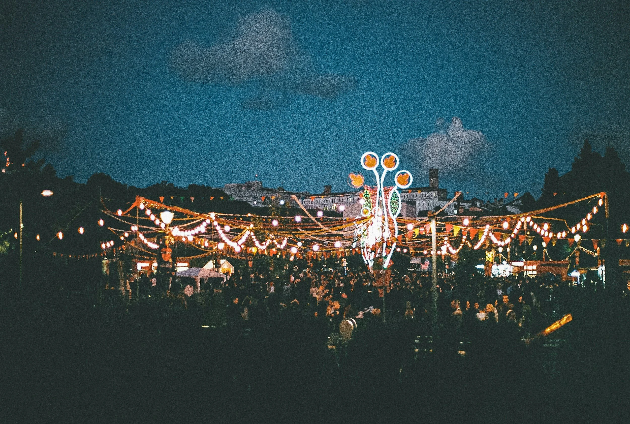 Nighttime scene of outdoor fair with string lights, decorations, and a large illuminated tree-like structure. Crowd of people visible, with buildings and trees in the background under a dark sky.