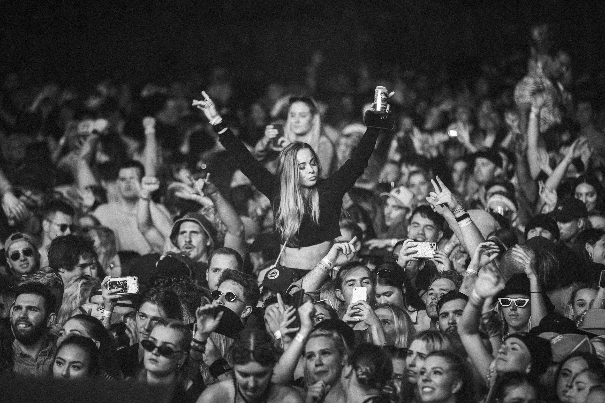 A large crowd of people at a concert or event, with a young woman standing on others' shoulders holding a drink and a phone, making peace signs with both hands.