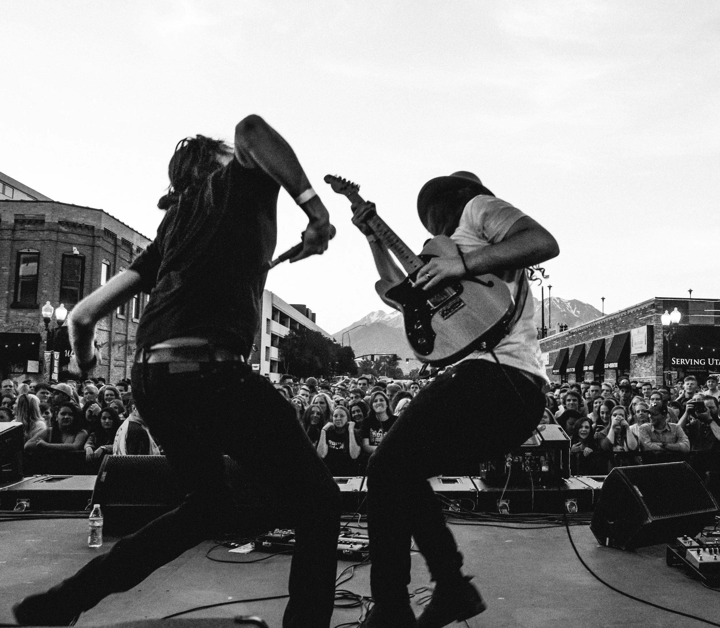 Black and white photo of two musicians performing on stage with a crowd watching, buildings, and mountains in the background.