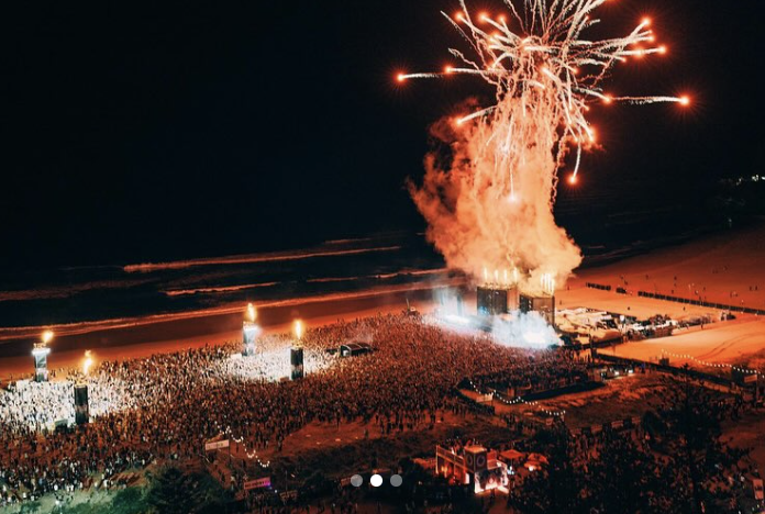 Fireworks exploding over a large crowd on a beach during night time with structures and illuminated area.