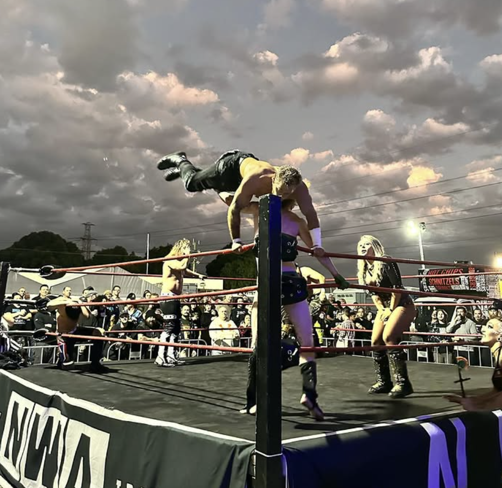 Wrestler performing a move off the top rope during a wrestling match in an outdoor arena, with spectators watching under a cloudy sky.